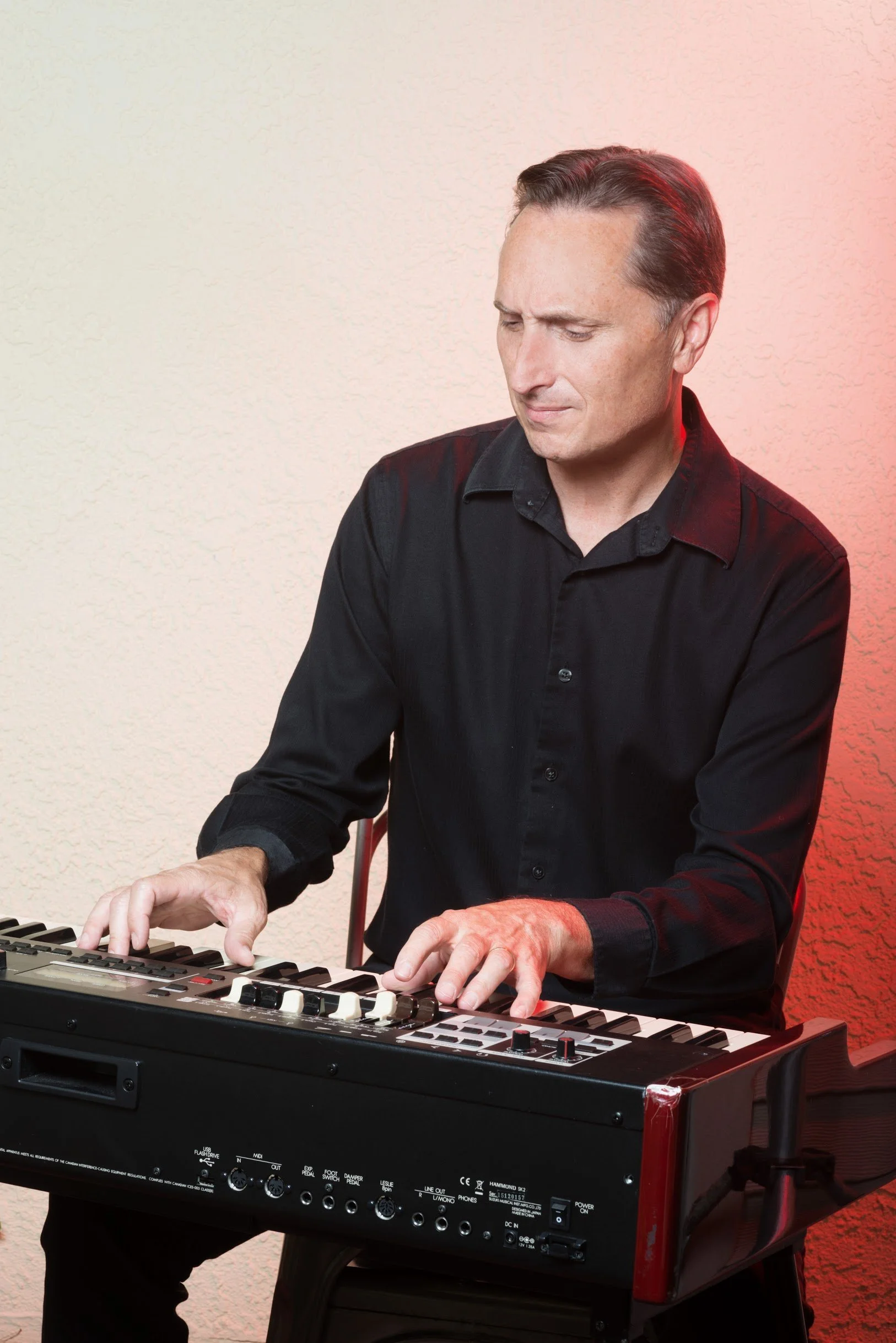 Man in a black shirt playing the keyboard on an ombre lit background.