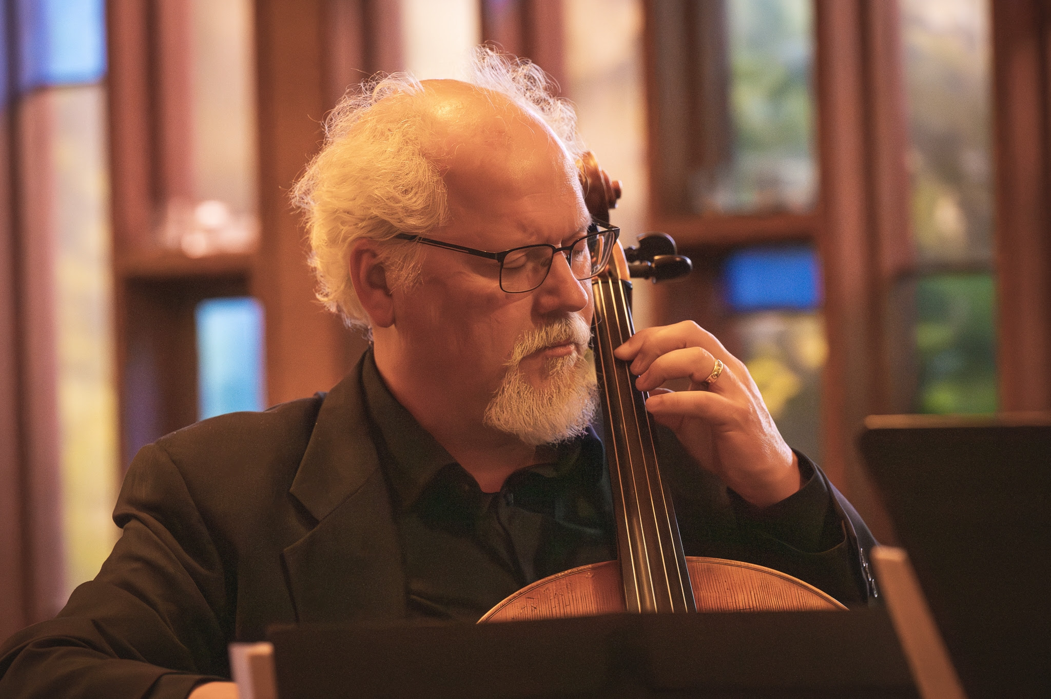 Man holding a cello, wearing a suit, with white hair and glasses in front of a row of stained glass windows.