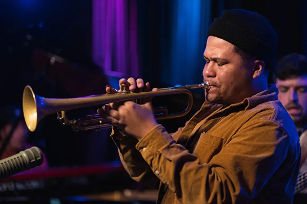 Man in a brown shirt with his eyes closed blowing a trumpet.