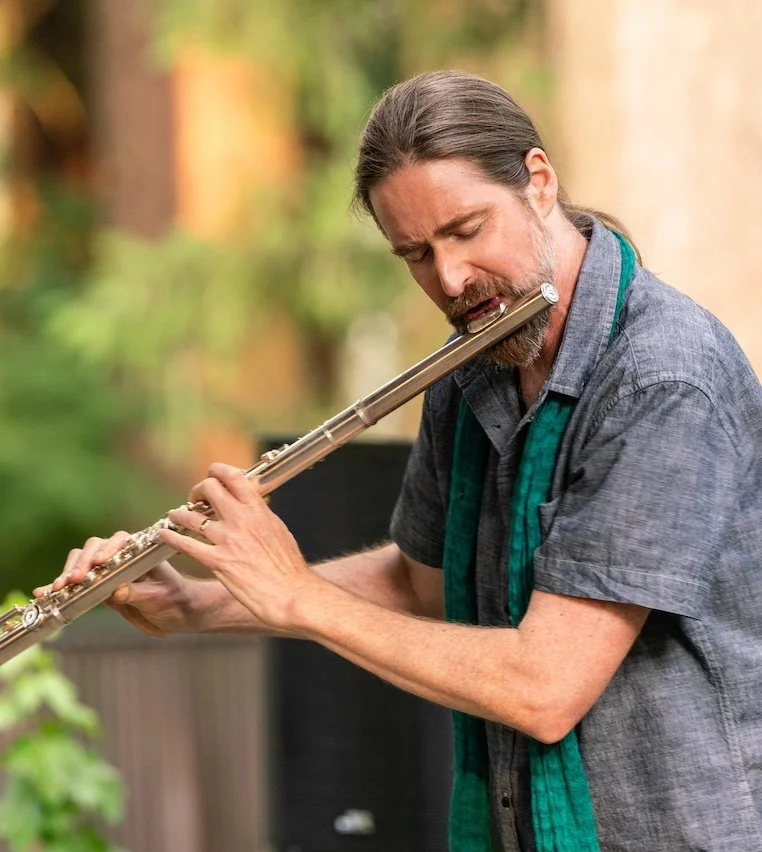 Man in a short sleeve shirt playing a metal flute with his eyes closed in an outdoor setting