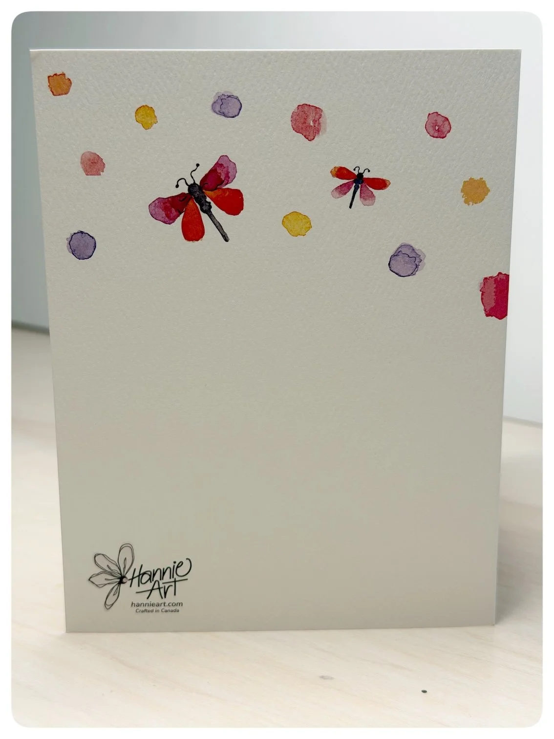 Back of surprise gift bow greeting card with watercolour dragonflies, colourful dots and Hannie Art logo