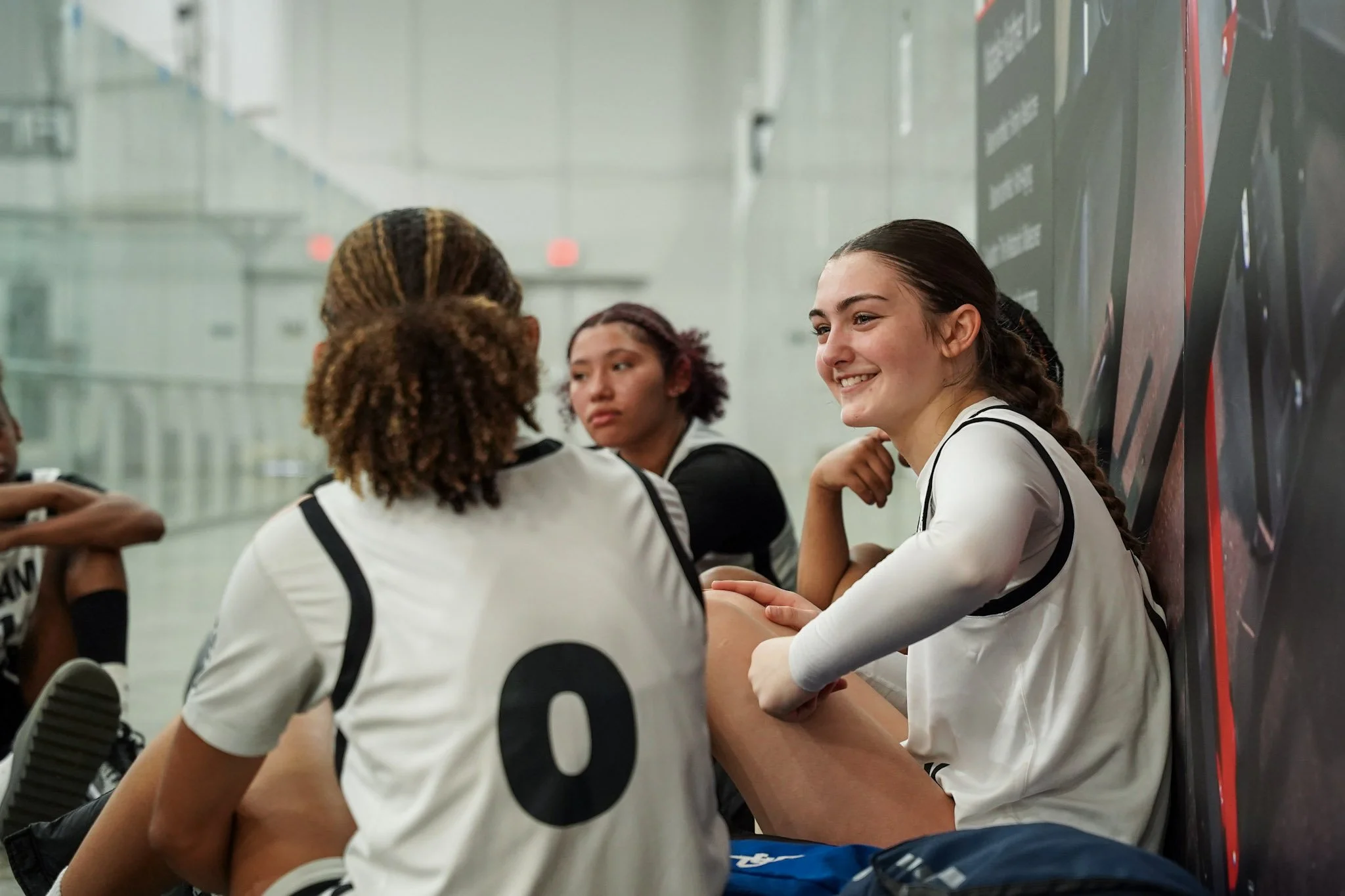 Young women athletes sitting and talking in a sports facility, wearing sports jerseys, with a smiling girl in the foreground.