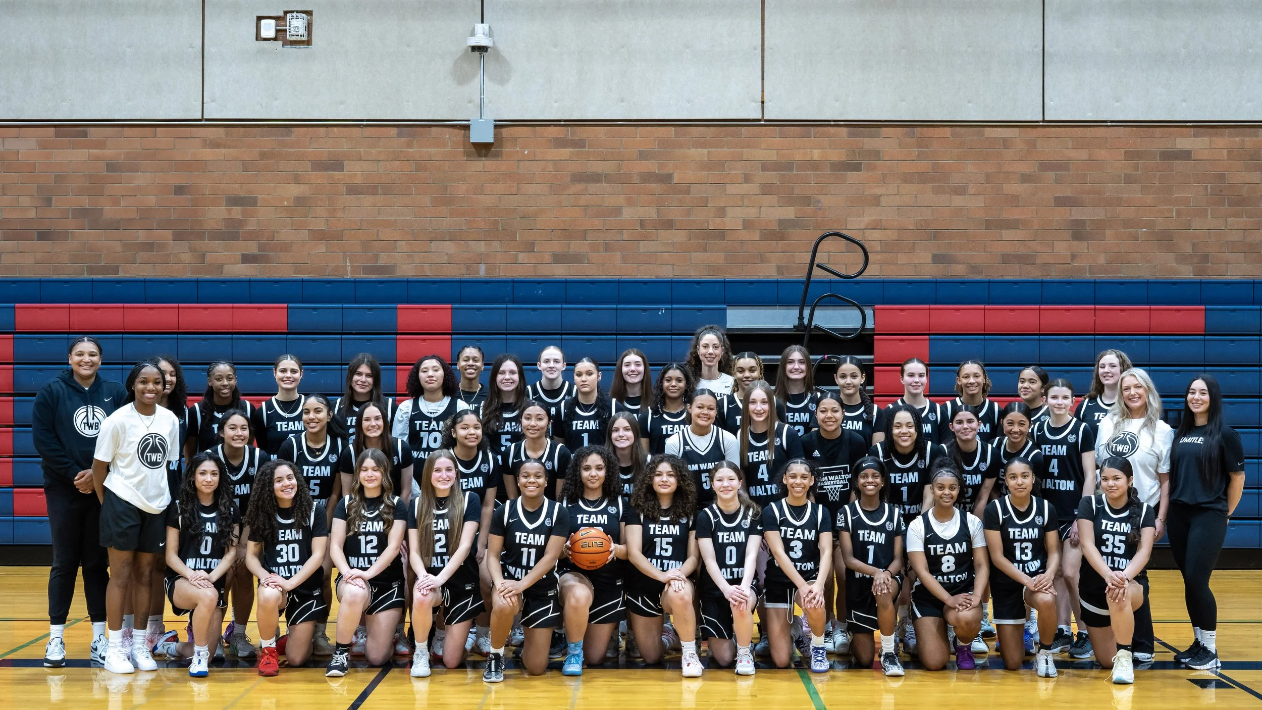 Group of young female basketball players and coaches posing for a team photo in a gymnasium with a basketball, wearing black and white uniforms with 'TEAM' and player numbers, behind the team is a blue and red gymnasium wall.