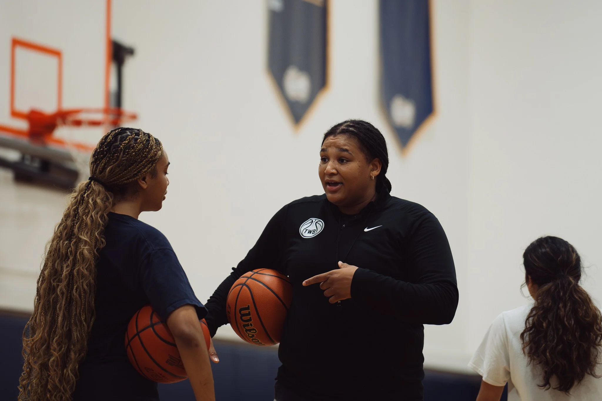 A female coach talking to a young girl basketball player in a gymnasium, with basketballs and a basketball hoop in the background.