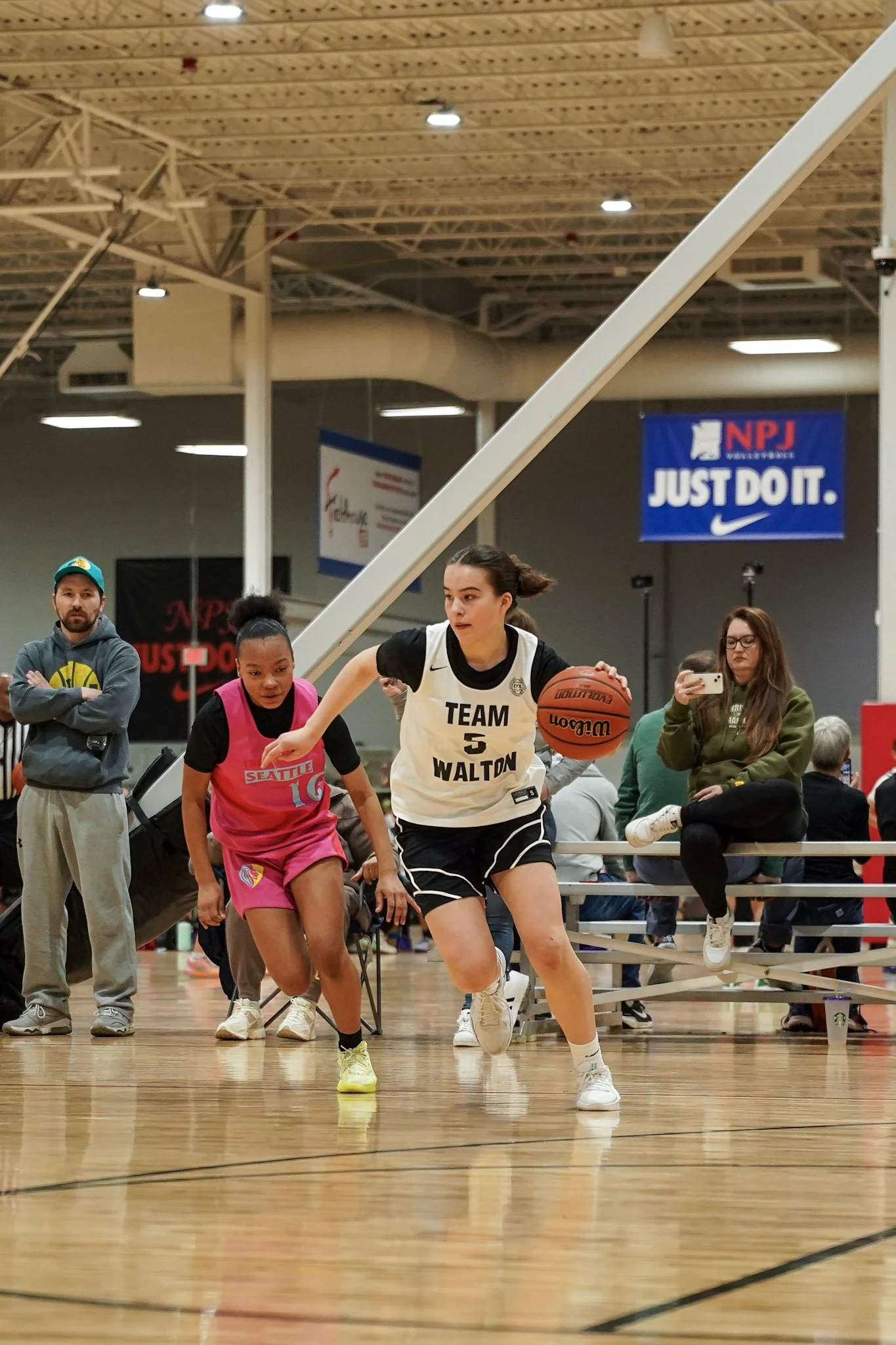 Two young female basketball players competing for the ball on an indoor basketball court, with one wearing a pink jersey and the other wearing a white jersey. Spectators and a coach are visible in the background.