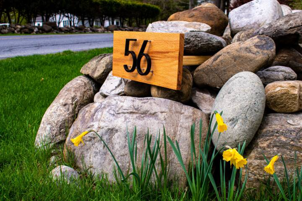 A wooden sign with the number 56 mounted on a stack of rocks, surrounded by daffodils and green grass by the roadside.