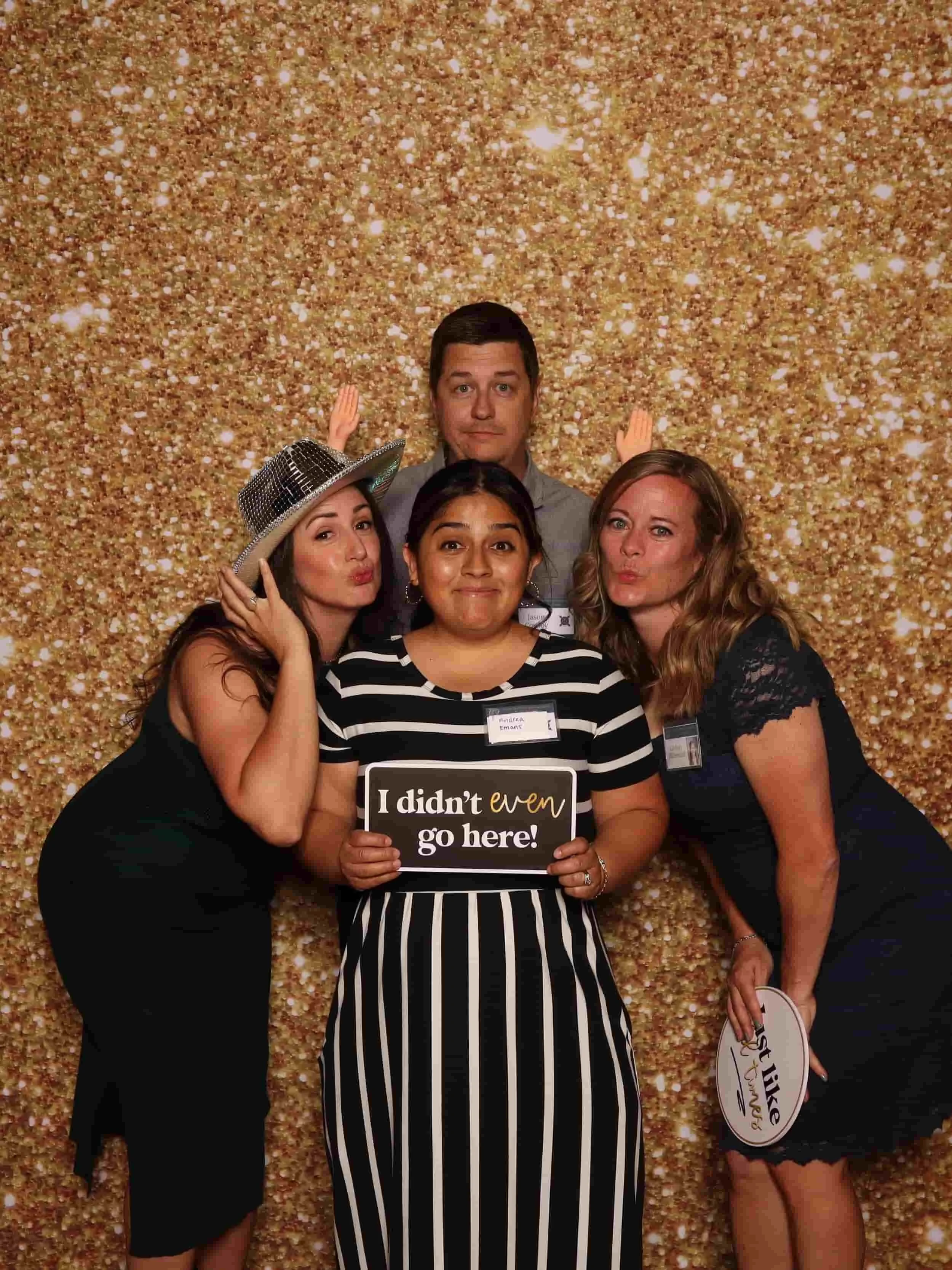 Four people posing in front of a glittery gold backdrop at a photo booth, two women on each side and a man behind them, with one woman holding a sign that says "I didn't even go here!" and another holding a sign that says "Just Biking." The women are