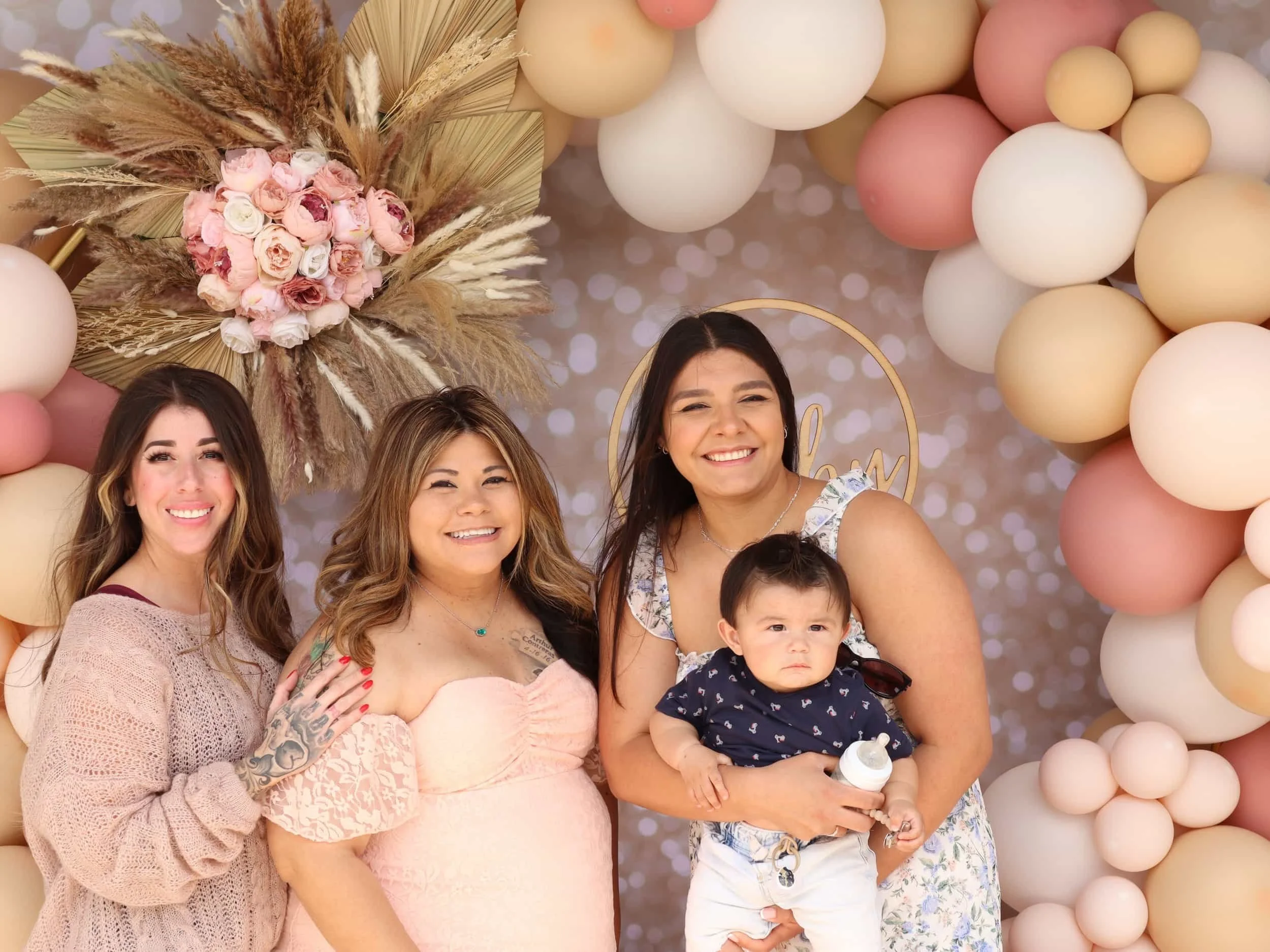 Four women, one baby, and pink and beige balloon decorations at a celebration. The women are smiling and standing together in front of a floral balloon arrangement with pampas grass and a pink rose bouquet, with a backdrop of blurred lights.