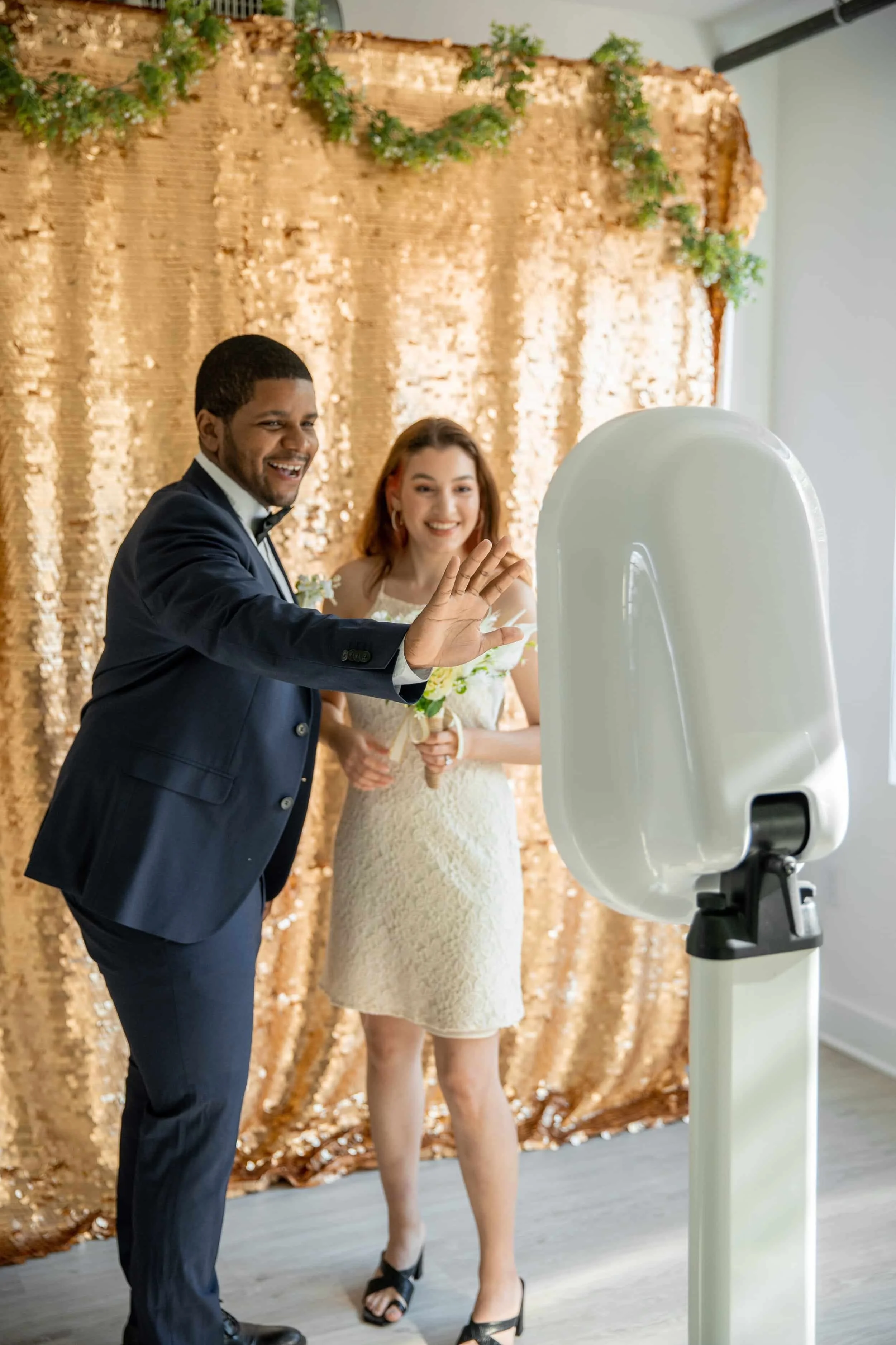 A couple dressed in formal attire taking a photo with a photo booth in front of a gold sequin backdrop. The man is in a navy suit and the woman is in a lace dress holding a bouquet of flowers.