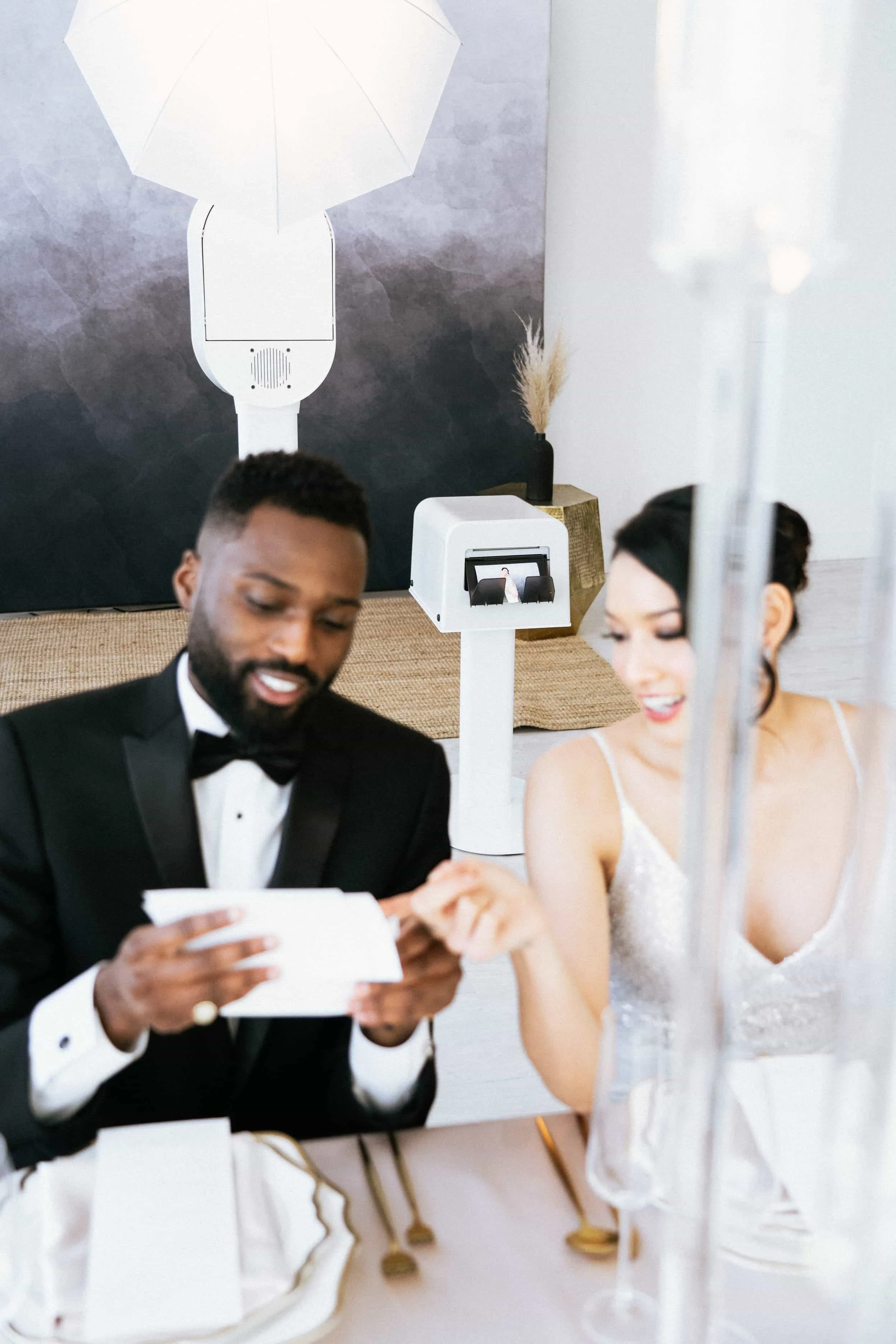A bride and groom dressed in wedding attire at a reception or event table, looking at a card or paper together, with a professional lighting setup in the background.