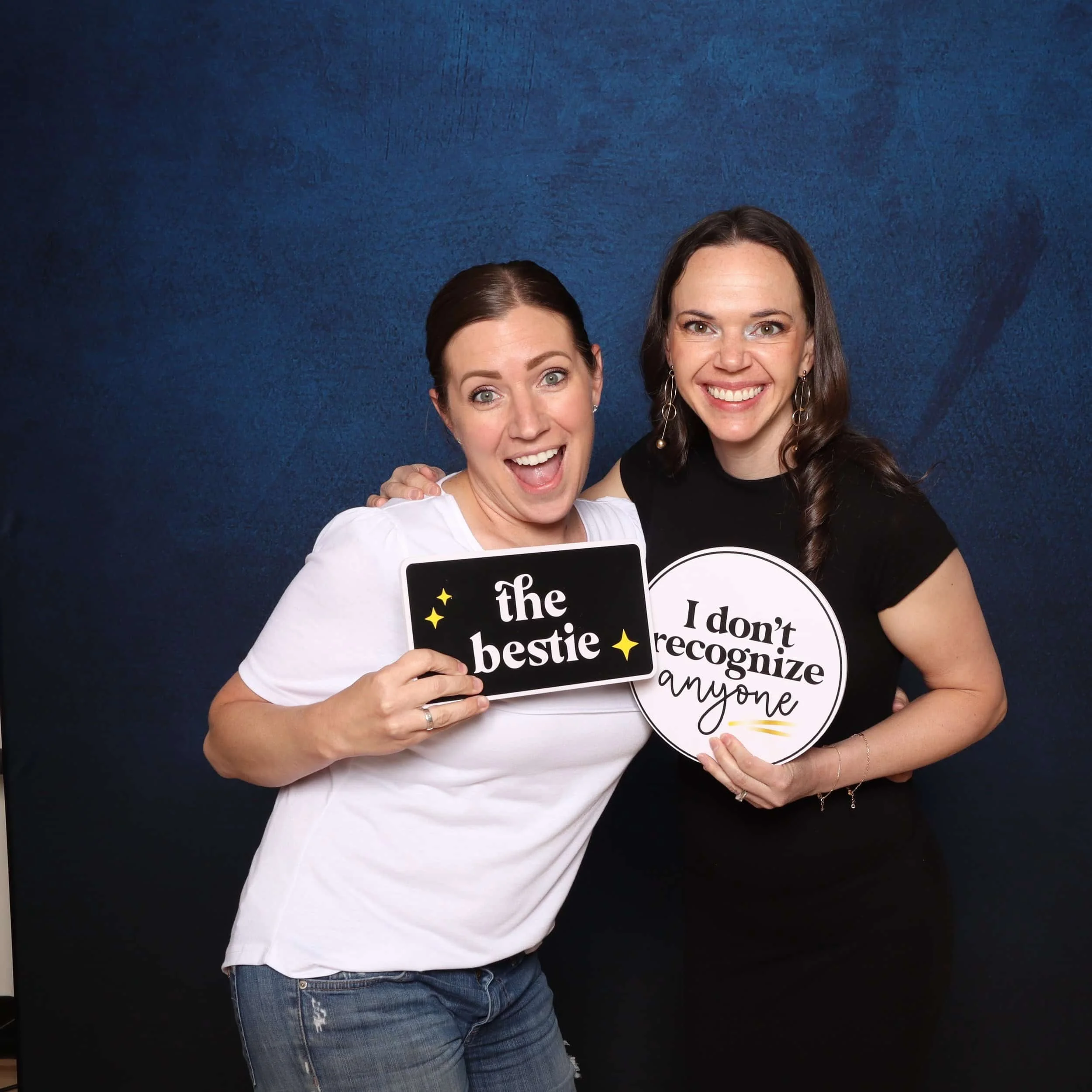 Two women smiling and holding signs in front of a blue background. The woman on the left holds a sign that says "the bestie" with star illustrations, and the woman on the right holds a sign that says "I don't recognize anyone."