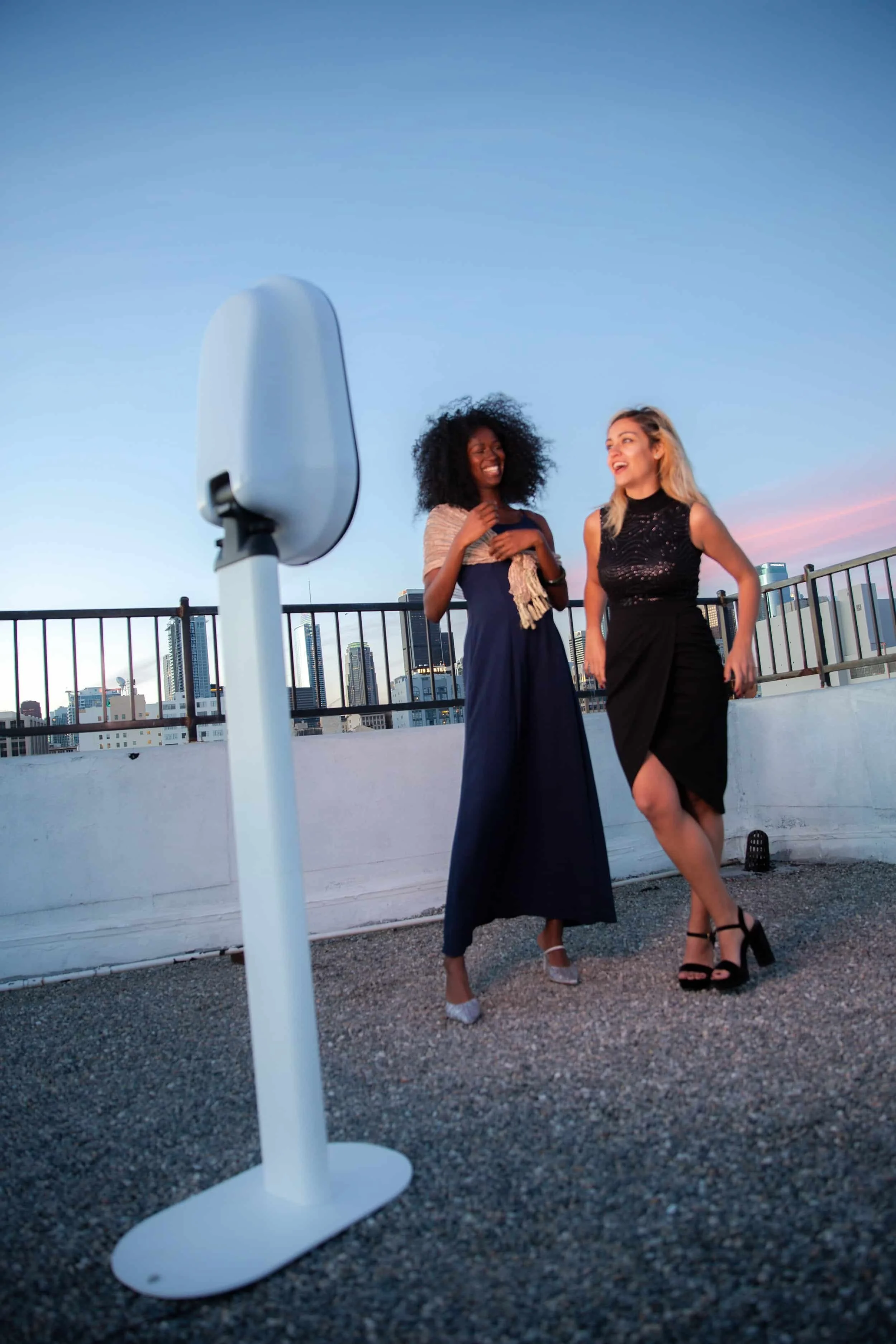 Two women standing on a rooftop, laughing and talking, with city buildings in the background, during sunset.