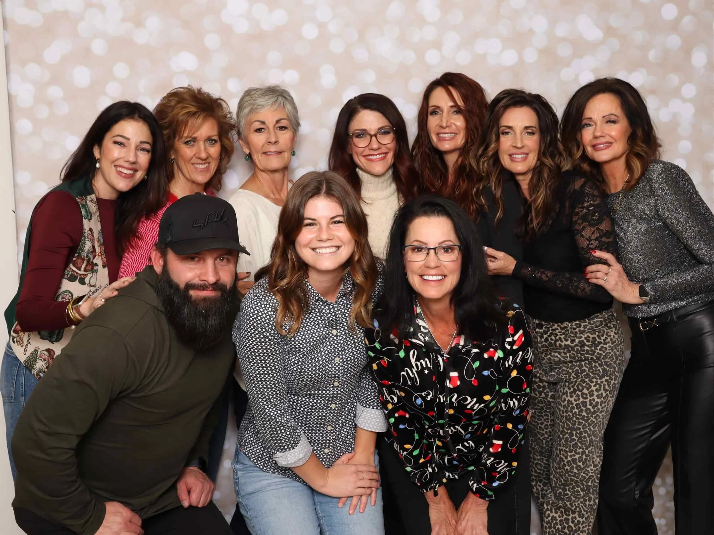 A group of twelve people, including men and women, posing together indoors in front of a light-colored, bokeh-style background. They are smiling and dressed in casual and festive clothing.