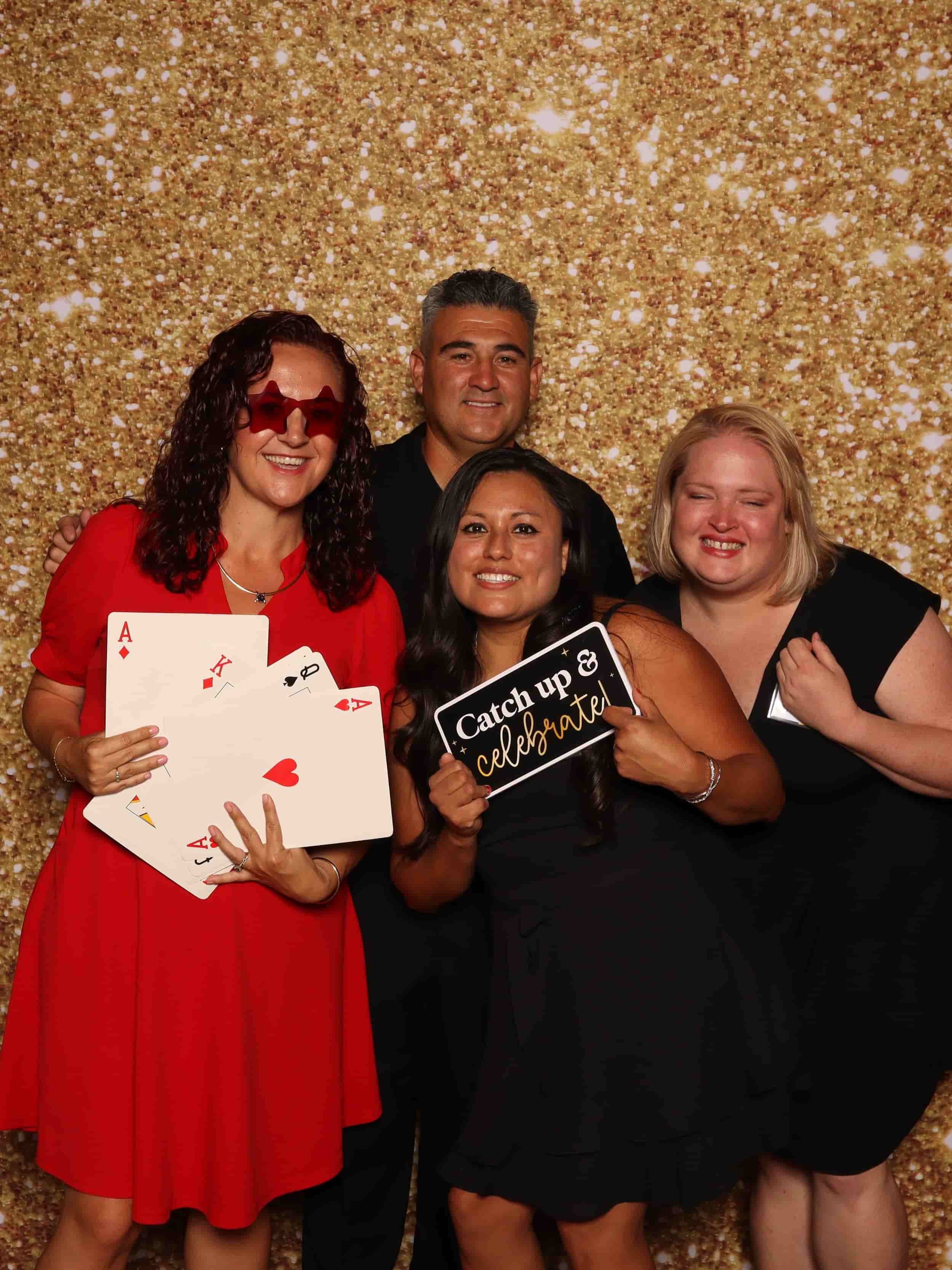 Four people celebrating at a party with a gold glitter background, holding playing cards and signs, smiling and dressed casually.