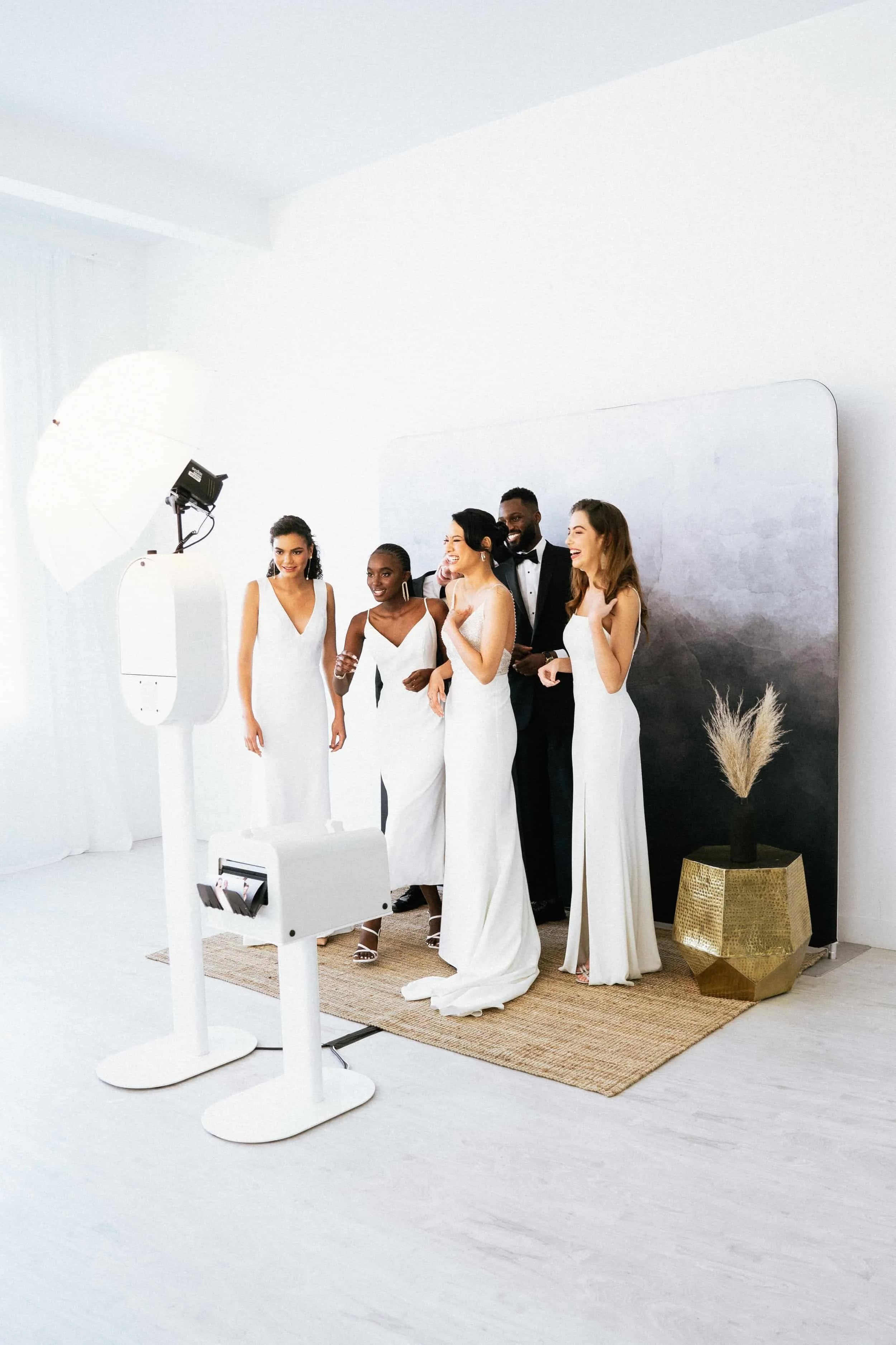 Group of six people dressed in formal attire enjoying a photo session in a white studio. Four women and two men, with the women in white dresses and the men in black tuxedos, smiling and laughing.