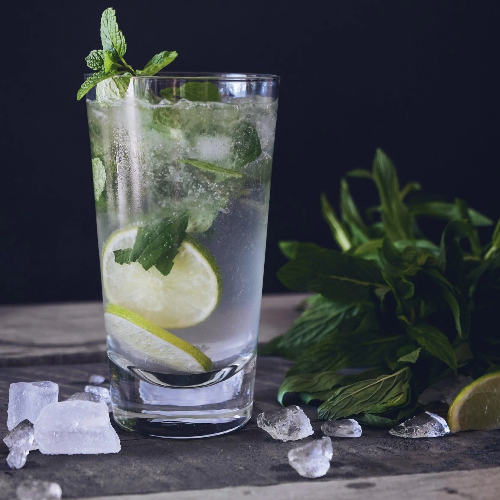 Ein Glas mit Limonade, Zitronenscheiben, Minzblättern und Eiswürfeln, daneben ein Bündel Minzblätter auf einem Holzbrett.