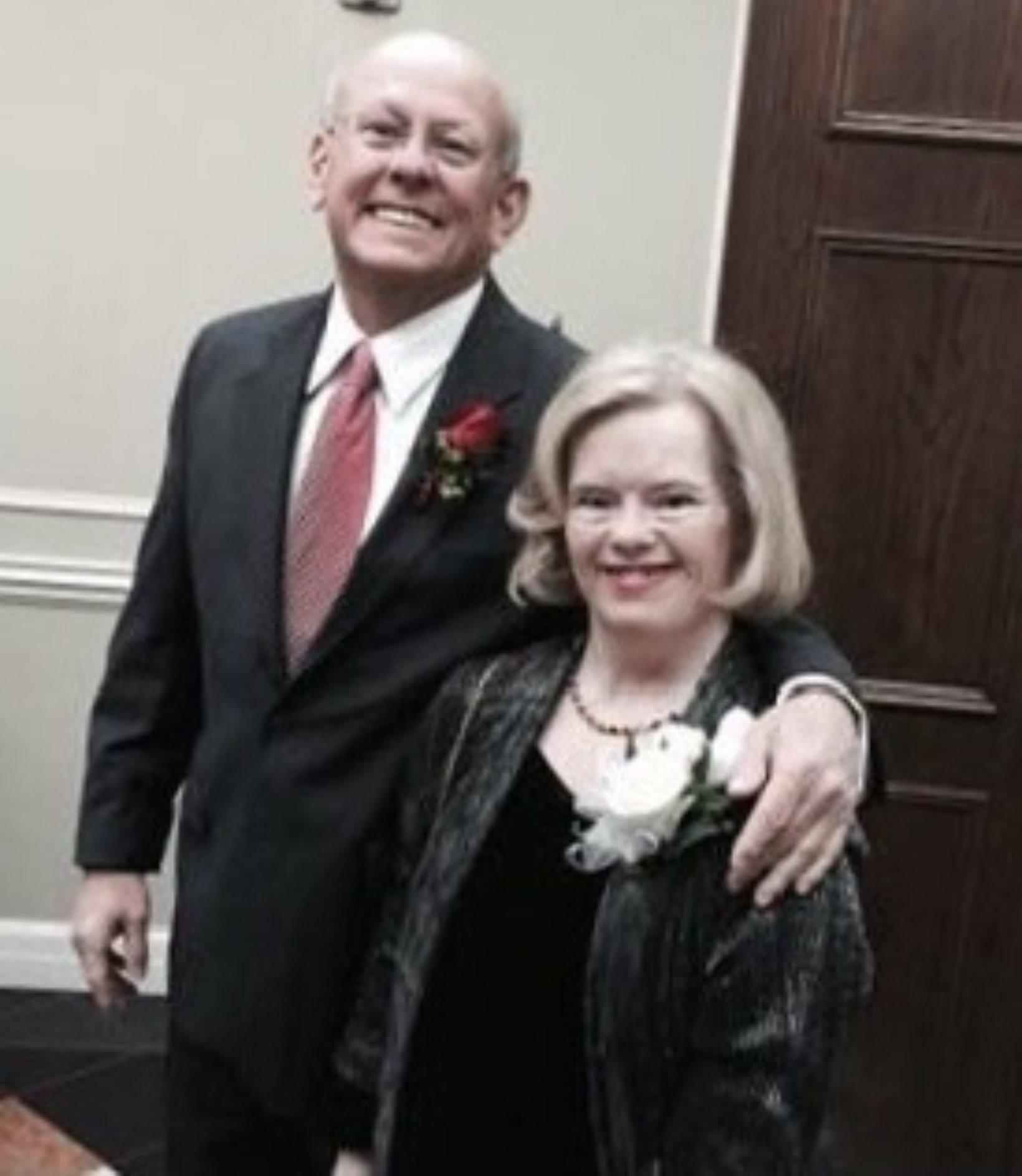 An elderly man and woman in formal attire, smiling for a photo. The man is in a suit with a red tie and a boutonniere, and the woman is in a black and silver dress with a floral corsage.