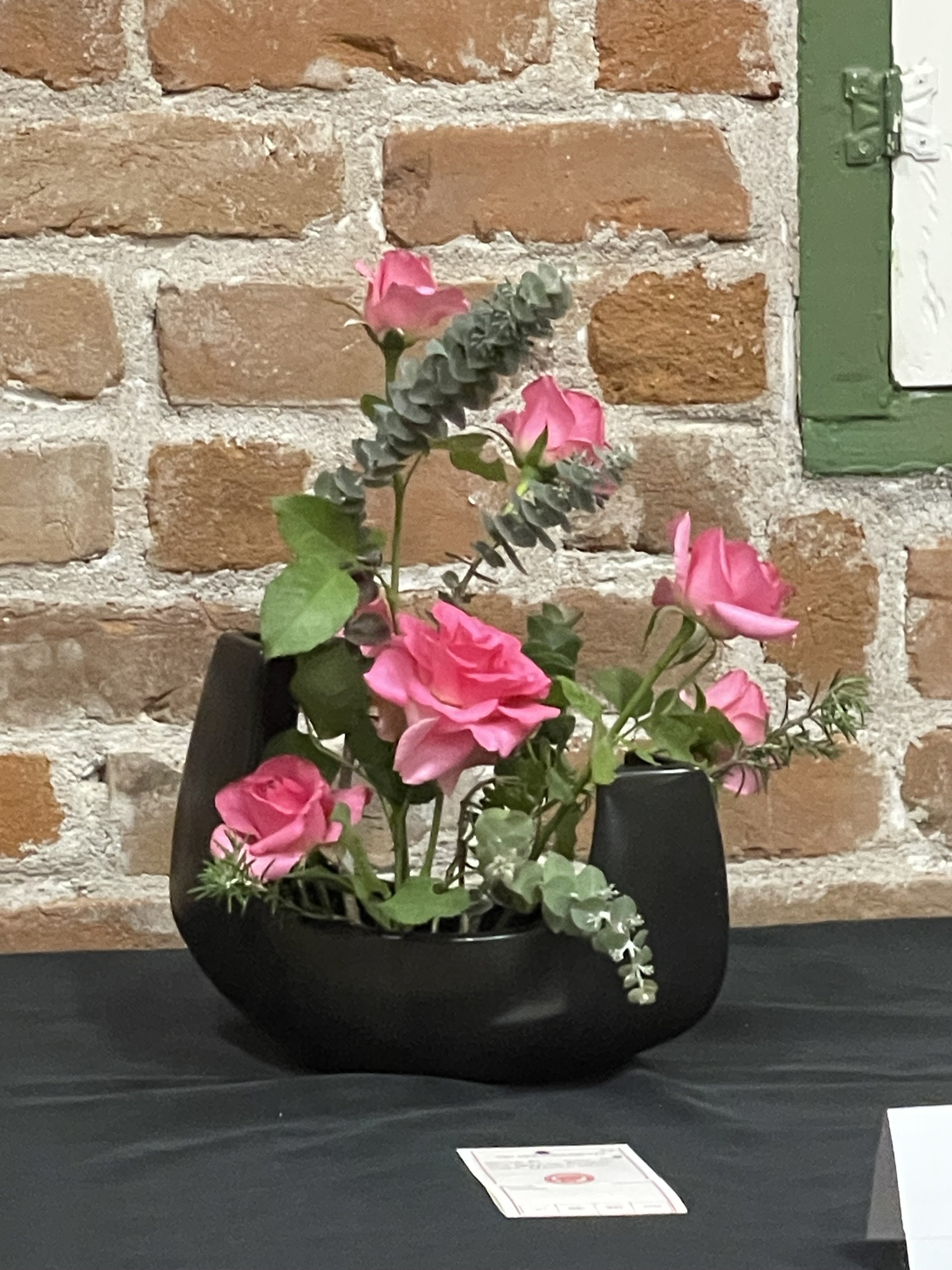 A black bowl filled with pink and white flowers, set on a black table with a brick wall in the background.