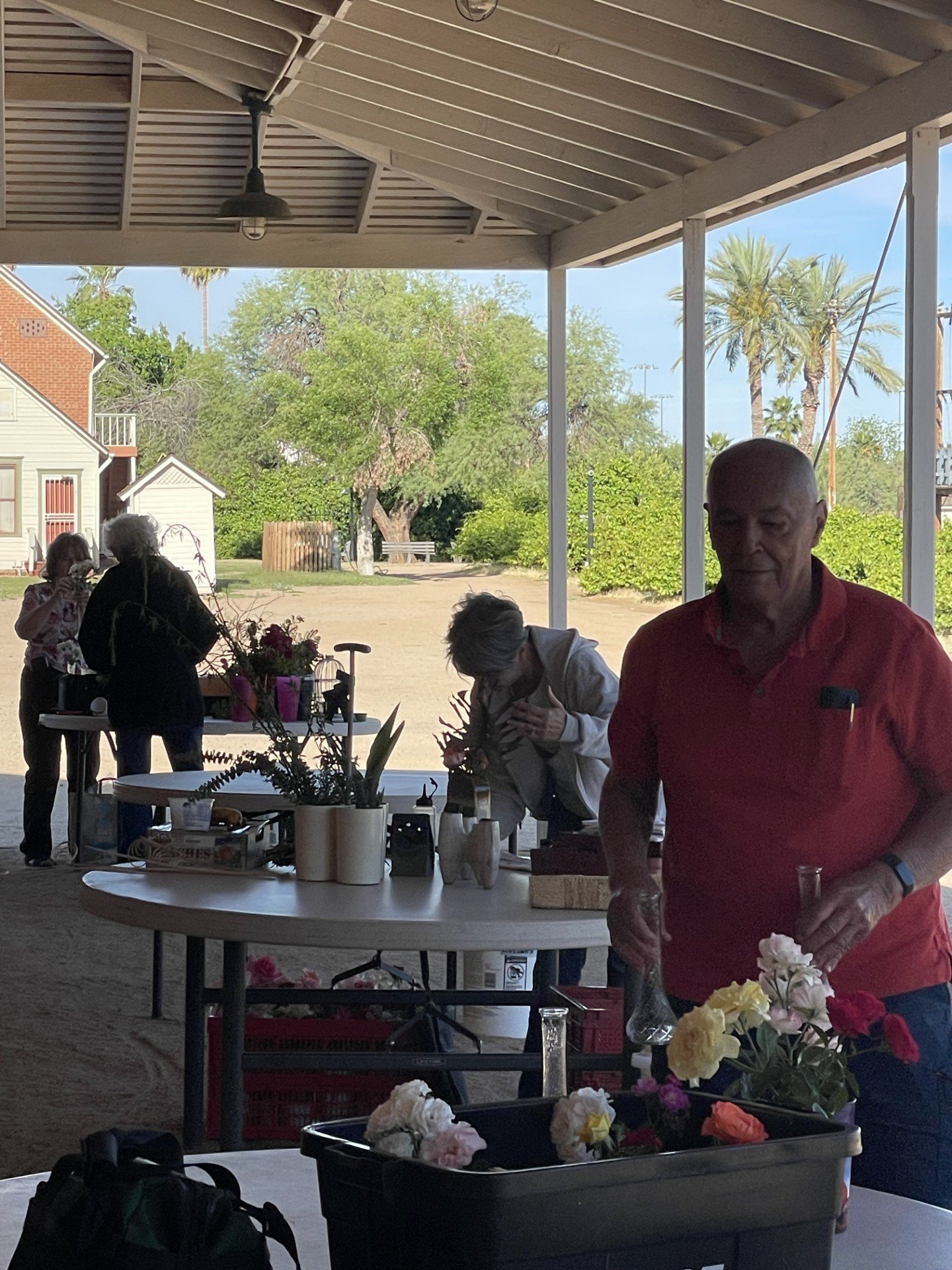 People gathering in a shaded outdoor area with trees and houses in the background, some preparing flowers on tables.