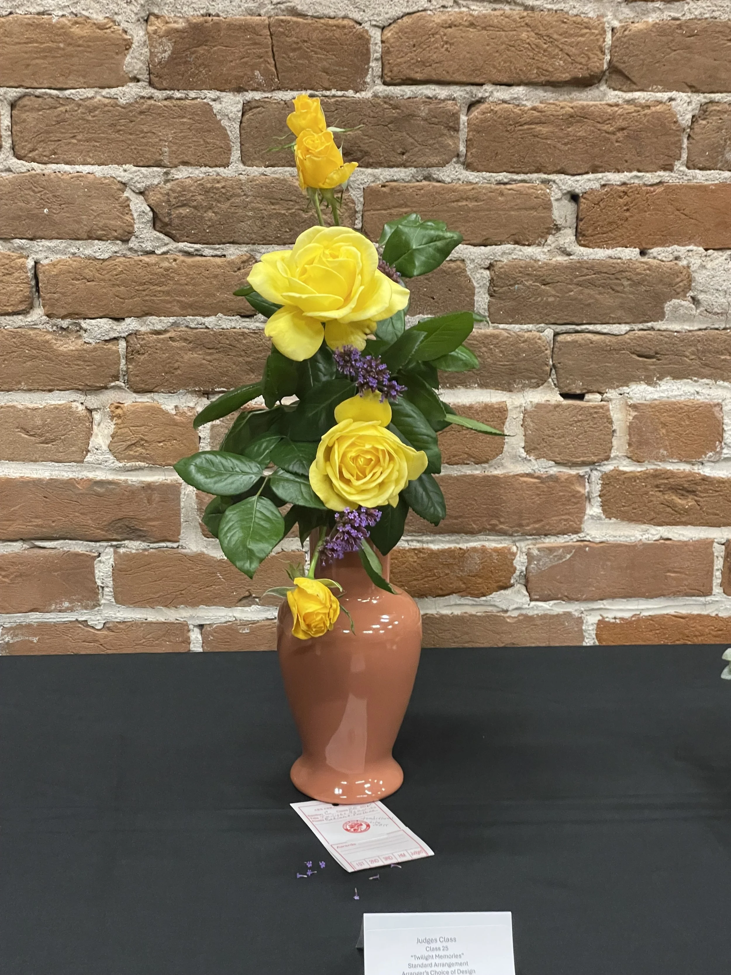 A floral arrangement with yellow roses and purple flowers in a pink ceramic vase on a black table against a brick wall background.