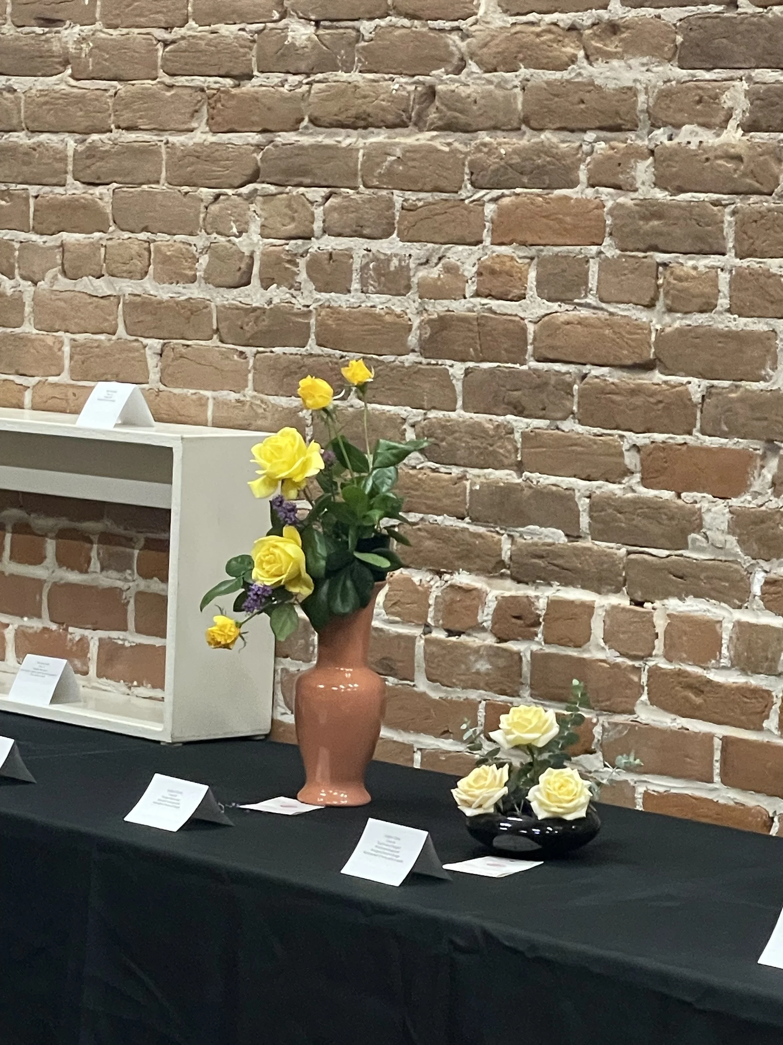 Display of various flower arrangements on a table with a brick wall background, including a clay vase with yellow roses and purple flowers, and a black vase with yellow roses, with informational cards in front of each arrangement.