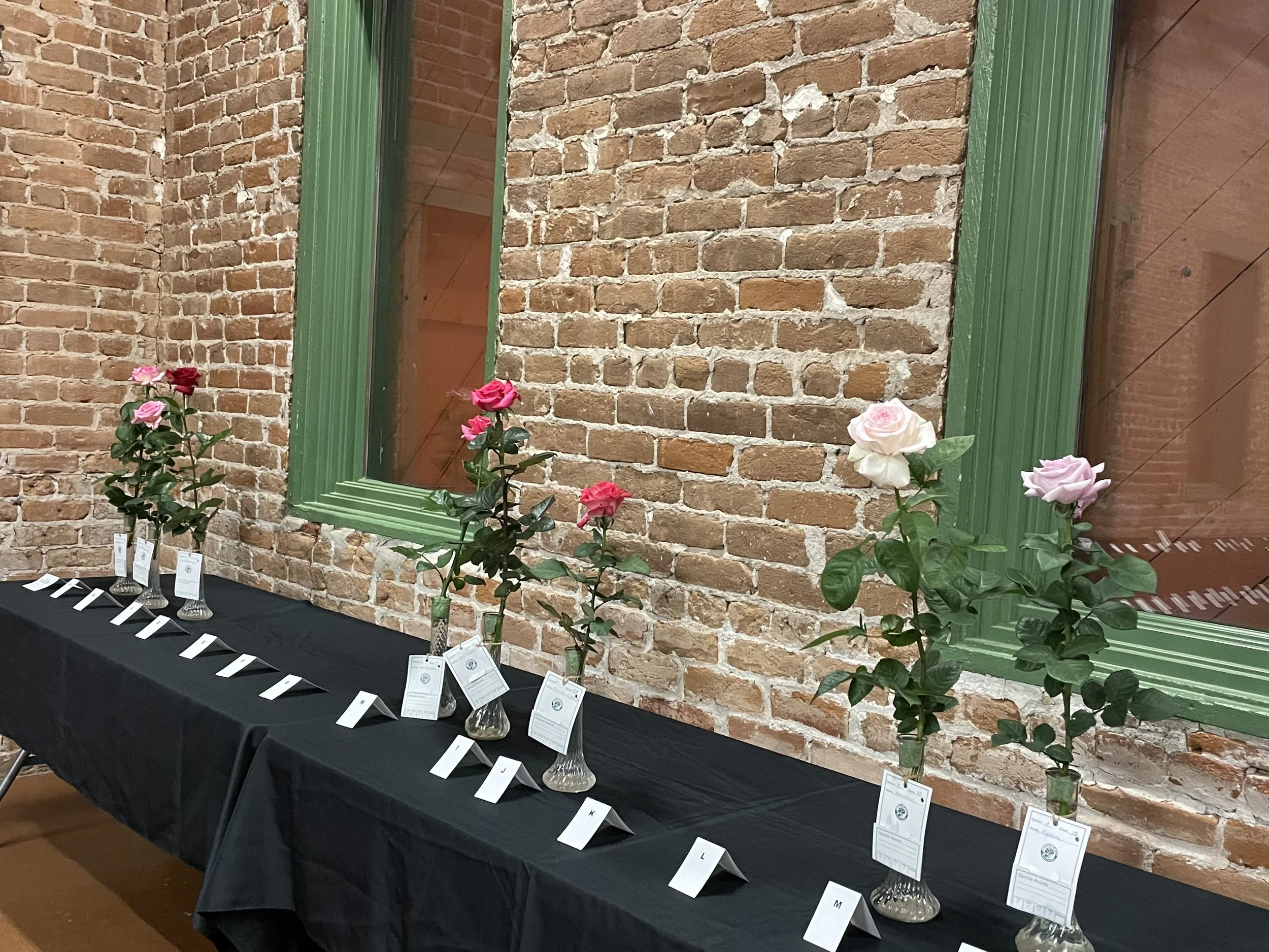A display of roses in vases arranged on a table with a black tablecloth, against an exposed brick wall with a green-framed mirror reflecting the interior.