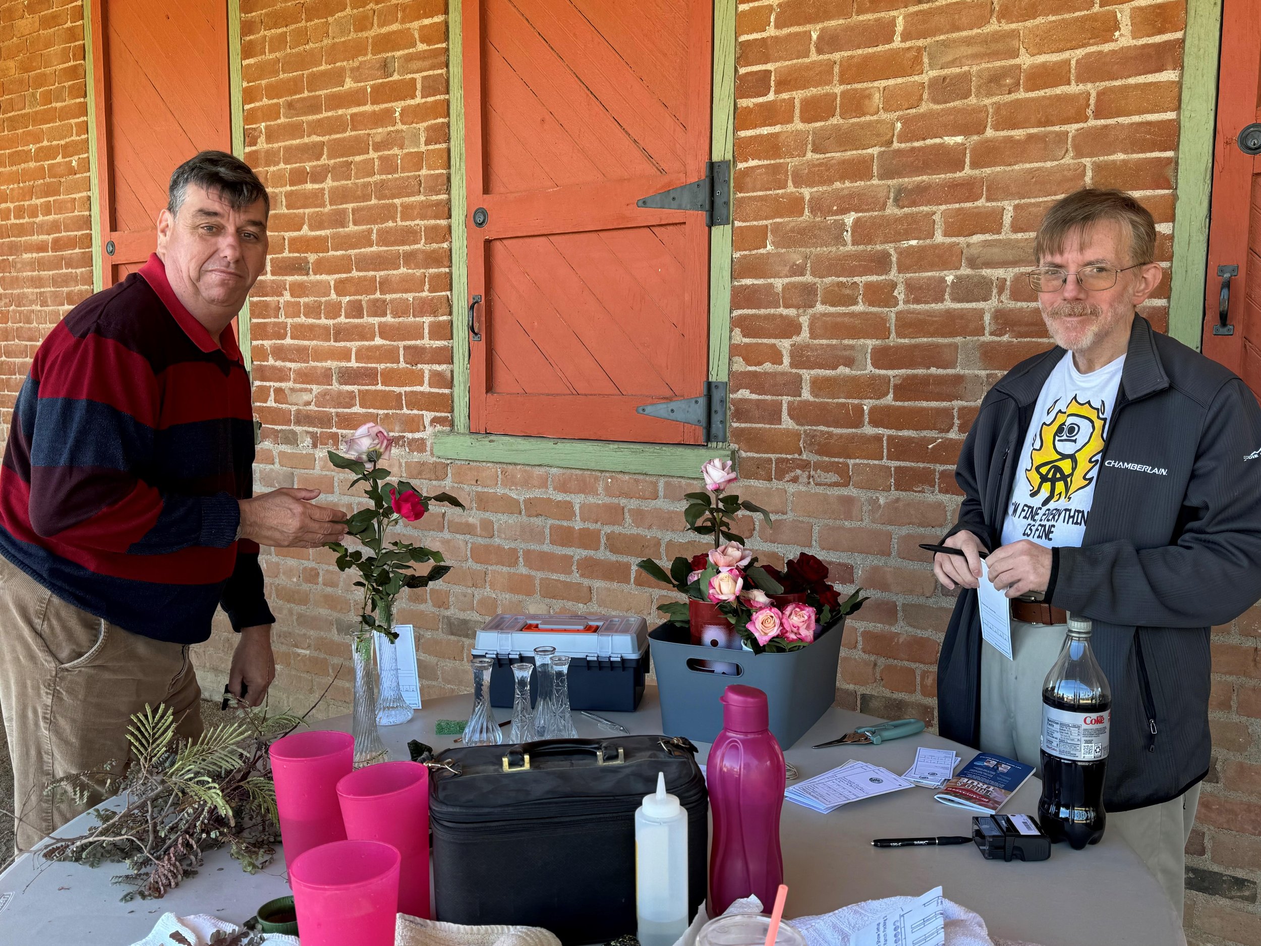 Two men standing behind a table with pink cups, vases, and flowers, in front of a brick wall with red shutters.