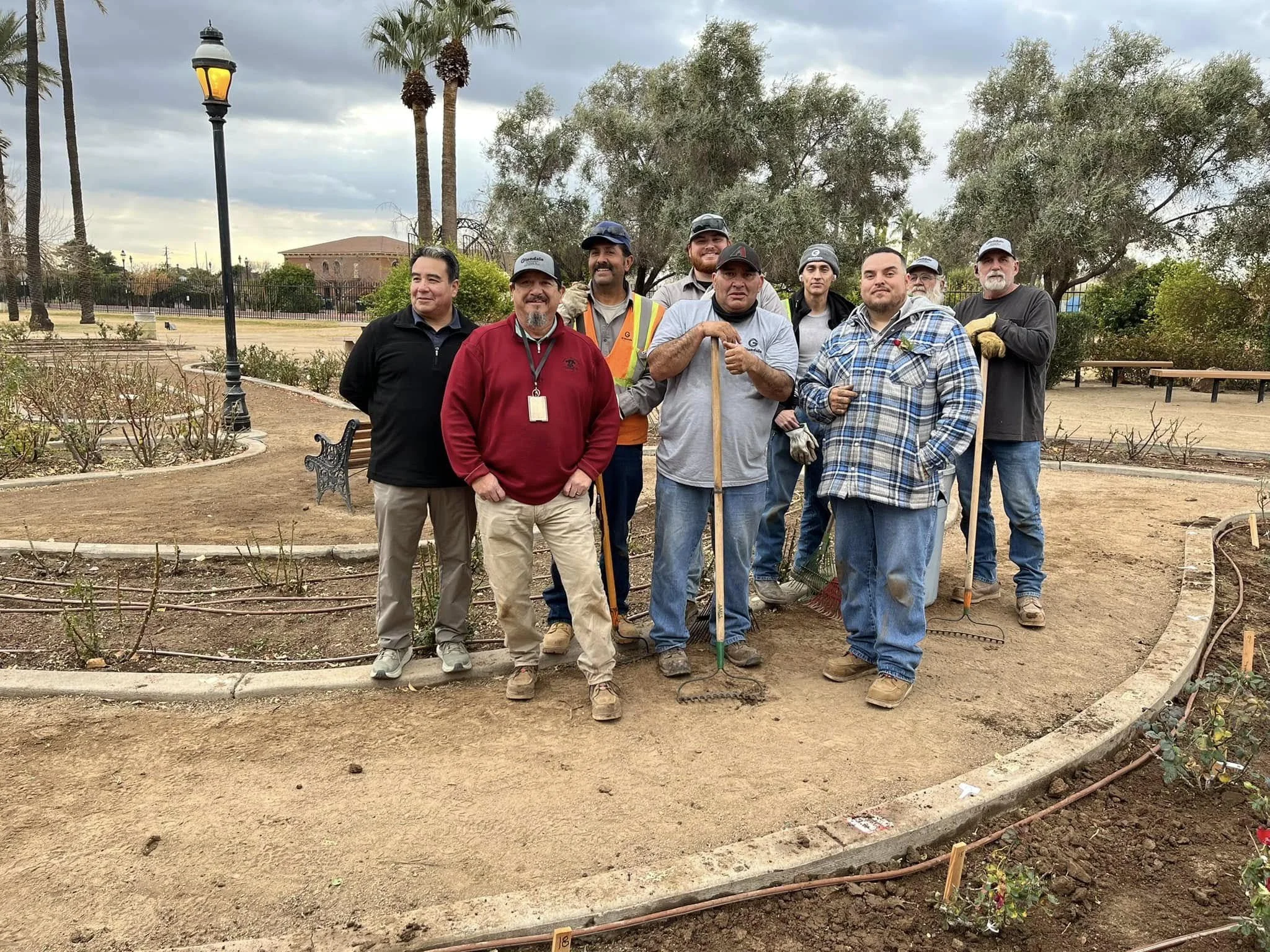 A group of ten men standing in a garden or park, some holding gardening tools like rakes and shovels. They are dressed casually, with some wearing hats and gloves. The background includes trees, a bench, a lamppost, and a fence. The sky is cloudy, indicating an overcast day.