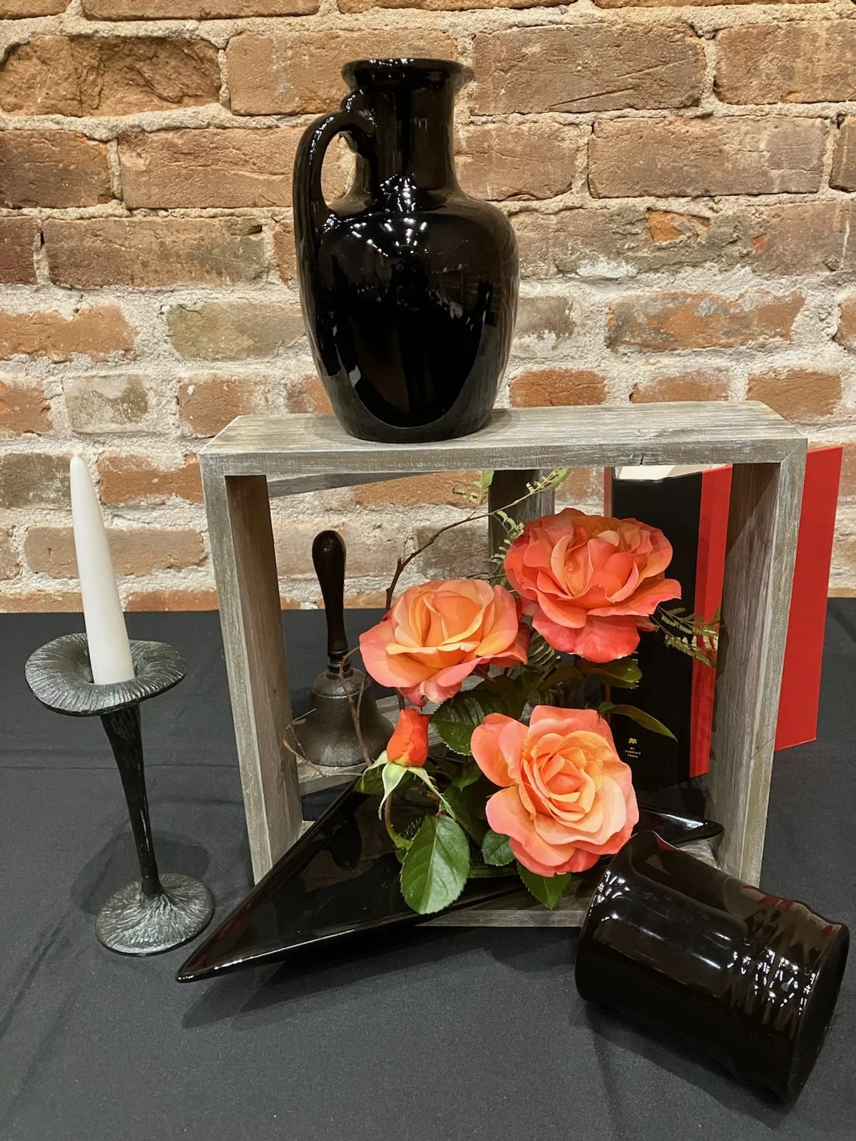 Decorative still life with a black ceramic vase on top, a tray with orange roses, a black plate, a black and white book, a black candleholder, a white tapered candle, a black candle, and a black glass piece, all set against a brick wall background.