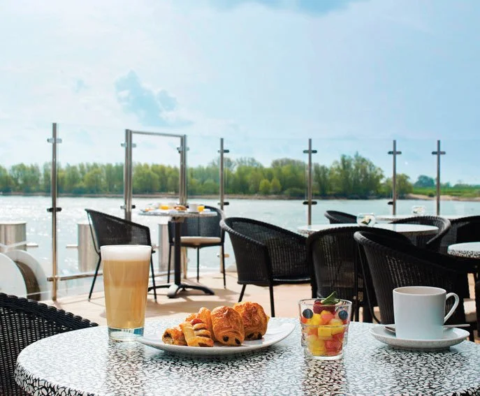 Breakfast table on a lakeside patio with coffee, fruit, and pastries, overlooking water and trees.