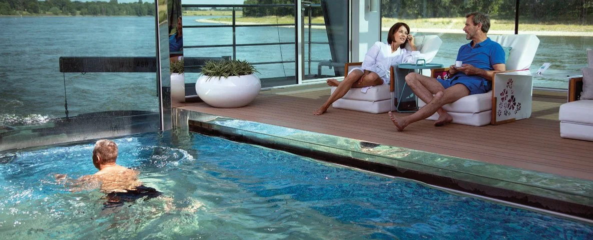 A man swimming in an indoor pool near a glass wall, with two women sitting on outdoor patio chairs by a lake, talking and relaxing. Emerald River Cruise.