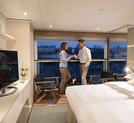 A woman and a man standing by a large window in a hotel room, clinking glasses of red wine and smiling at each other. On River Cruise