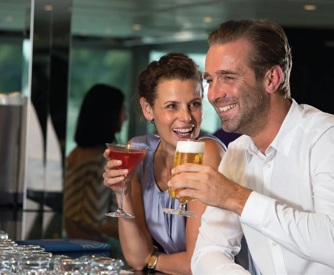 A man and woman smiling at each other while sitting at a bar, each holding a drink. The woman has a cocktail, and the man has a beer.