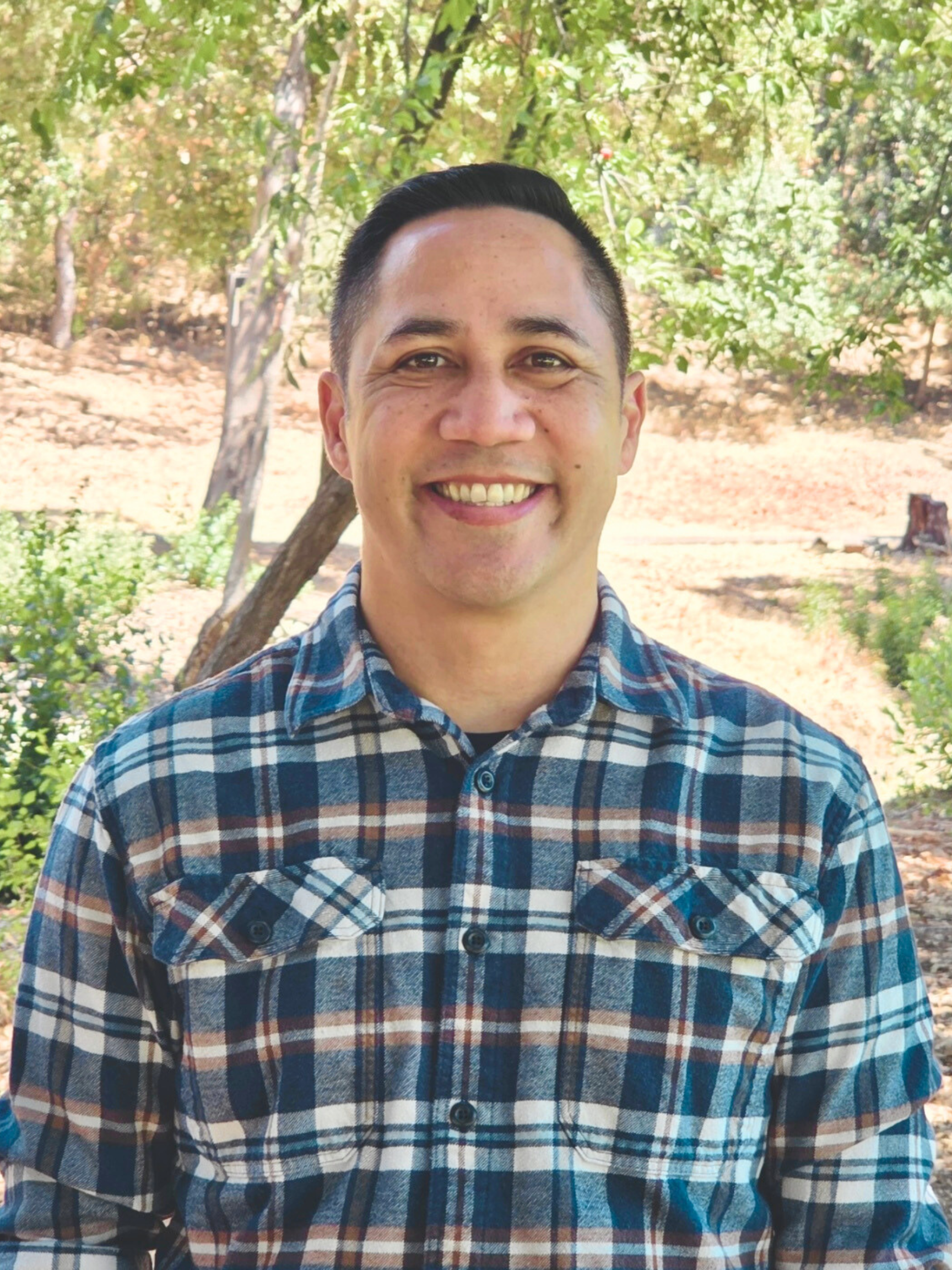 A smiling man standing outdoors in a natural setting with trees and greenery.