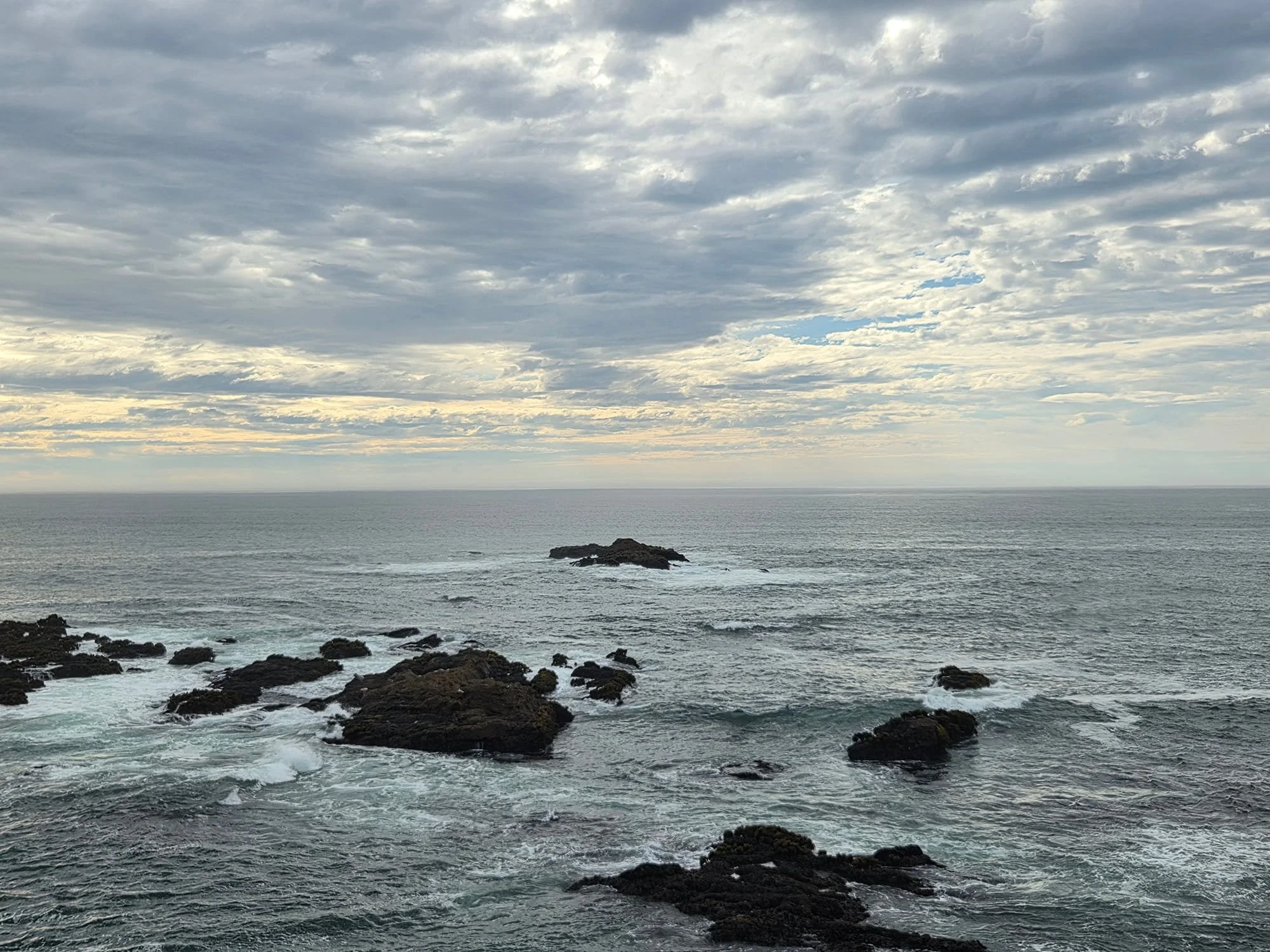 Overcast sky over a rocky ocean shoreline with waves crashing against rocks.