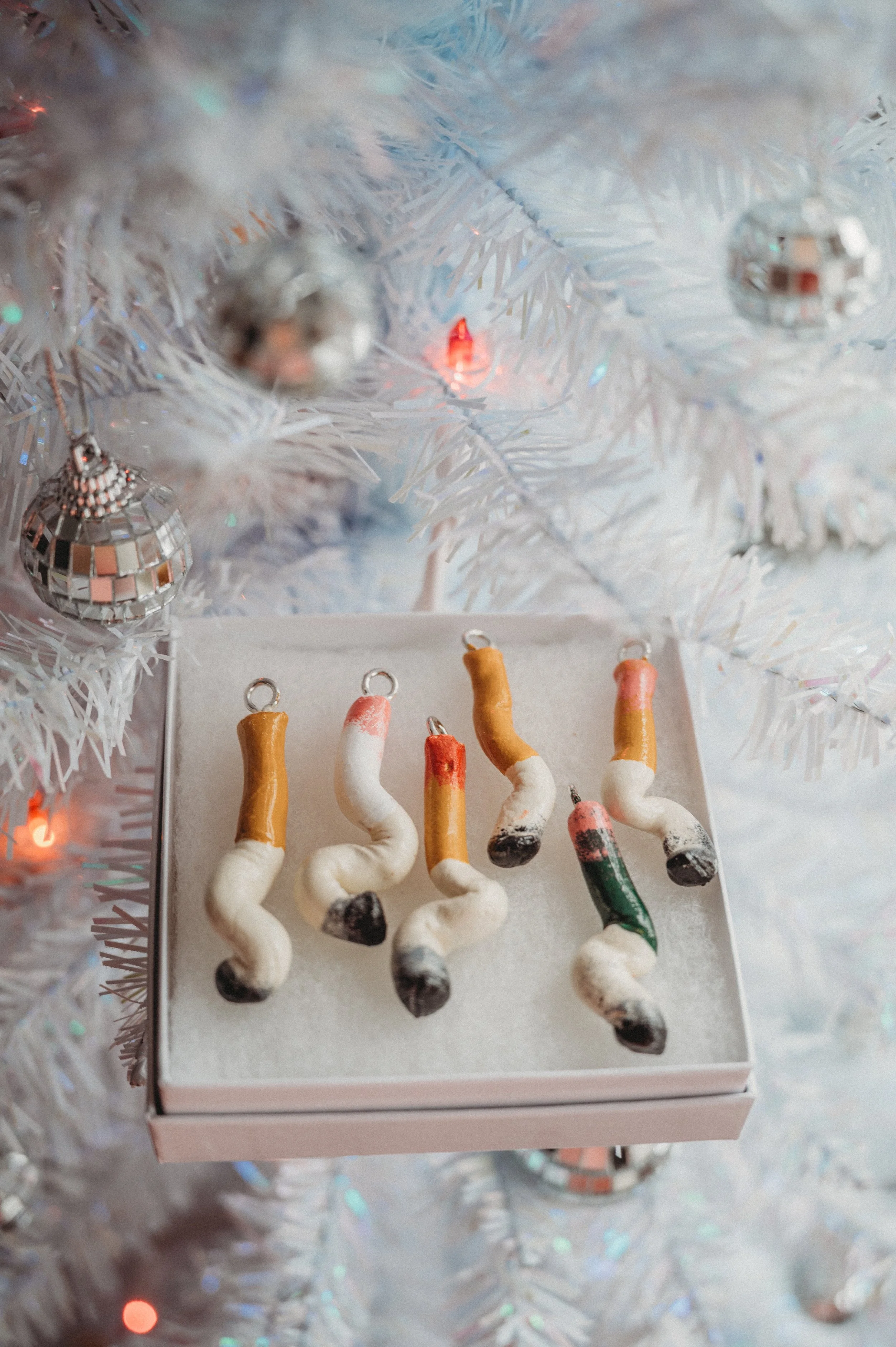 Set of six cigarette ornaments in various colors, placed on a white box under a white Christmas tree with silver ornaments and multicolored lights.