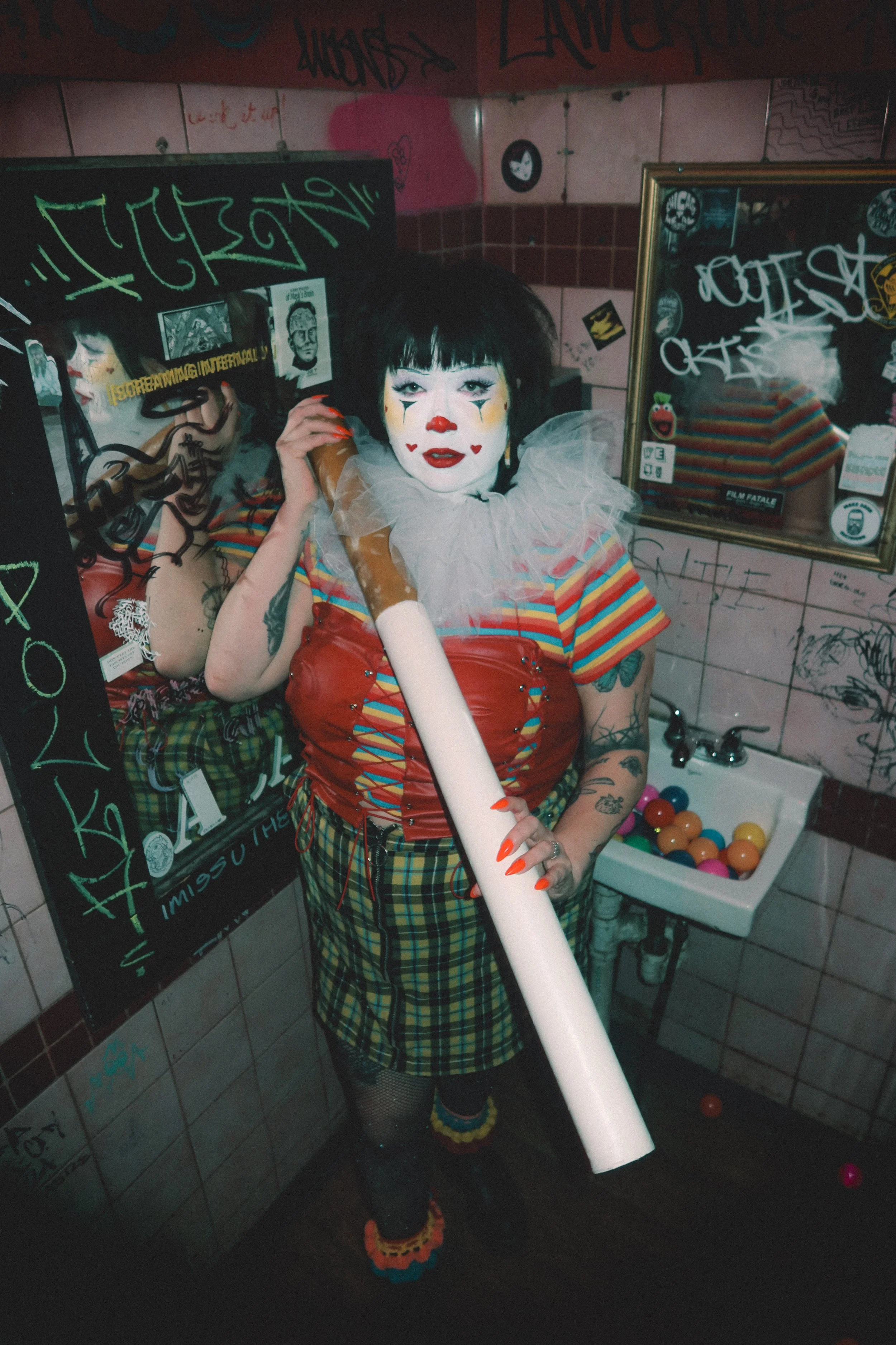Person with clown makeup and colorful attire holding a large fake cigarette in a graffiti-covered bathroom.