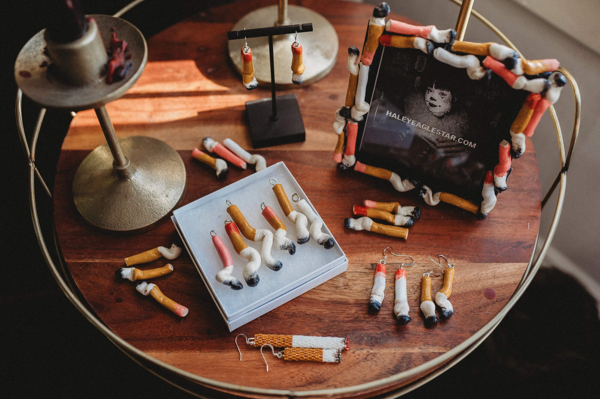 Collection of jewelry and accessories on a wooden table, including earrings, a picture frame decorated with cigarette-shaped jewelry, and display stands.
