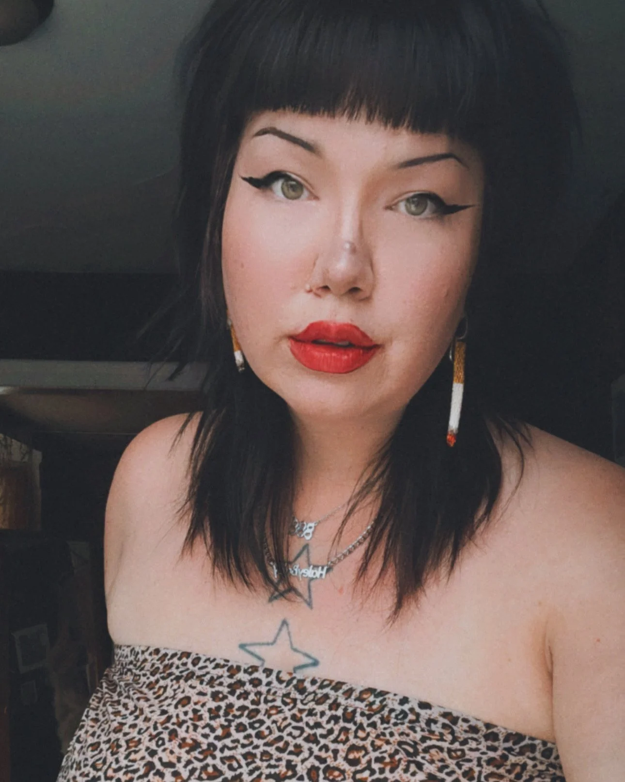 Person with black hair and bangs, wearing leopard print clothing, red lipstick, cat eye makeup, star tattoo on chest, and earrings in a dimly lit room.