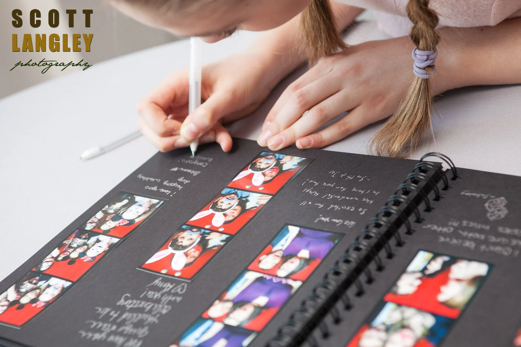 A young girl with braided hair using a white pen to write in a scrapbook filled with photo booth pictures and handwritten notes on a gray surface.