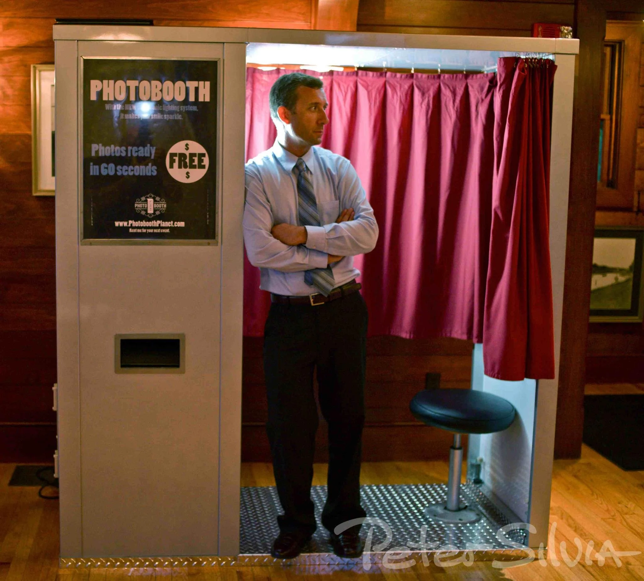 A man in business attire standing inside a photo booth with red curtains. A sign on the booth advertises free photos ready in 60 seconds.