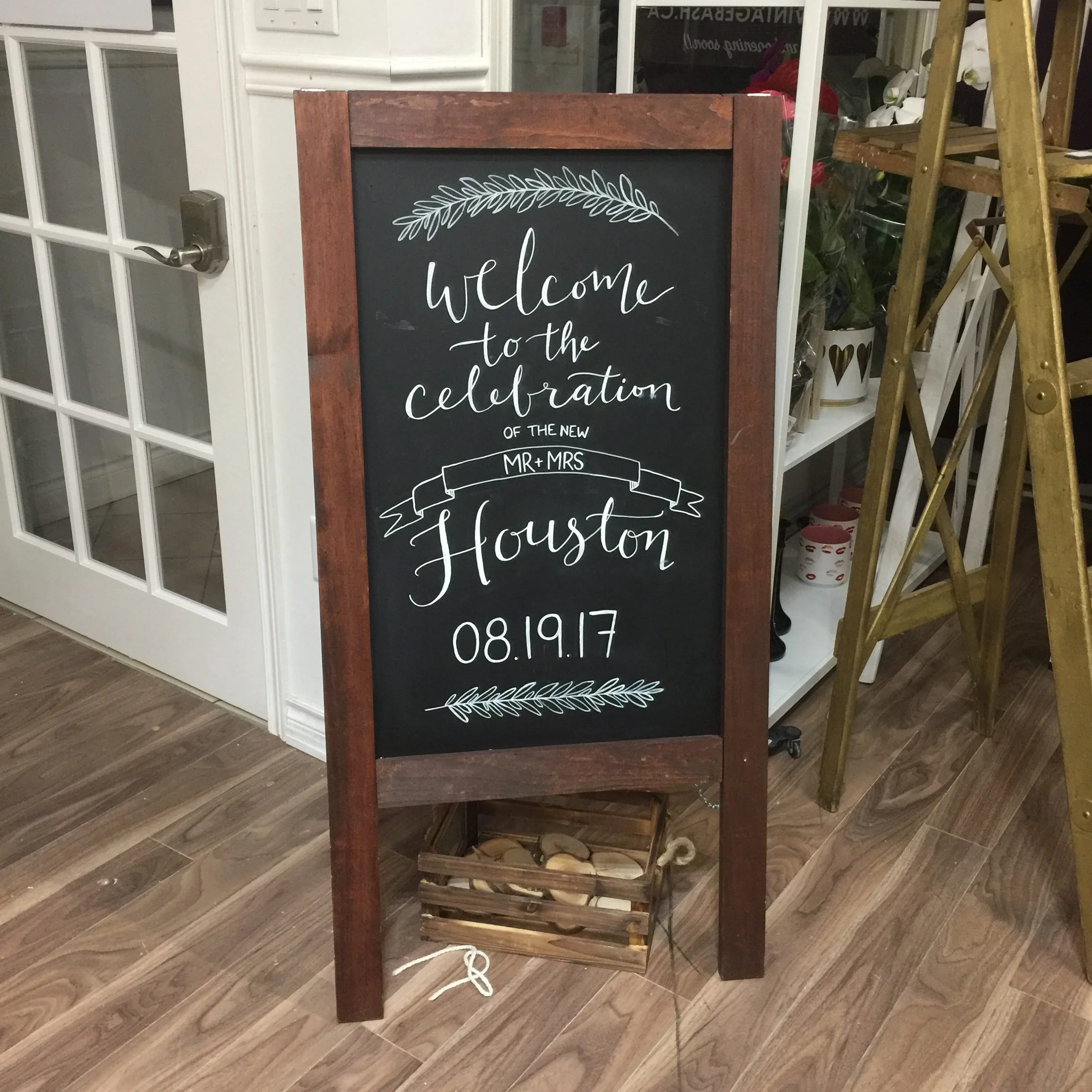 A decorative chalkboard sign with wooden frame announcing a celebration of the wedding of Mr. and Mrs. Houston on August 19, 2017, with floral designs at the top and bottom.