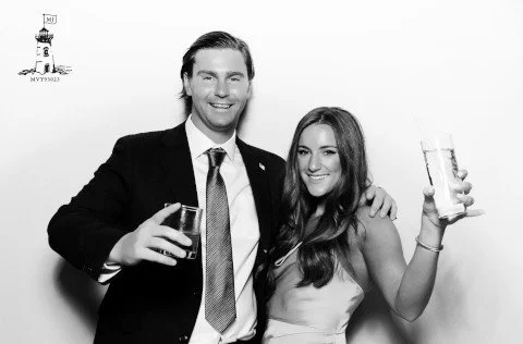 A man and a woman smiling and holding drinks at a social event, standing close together in front of a plain background.