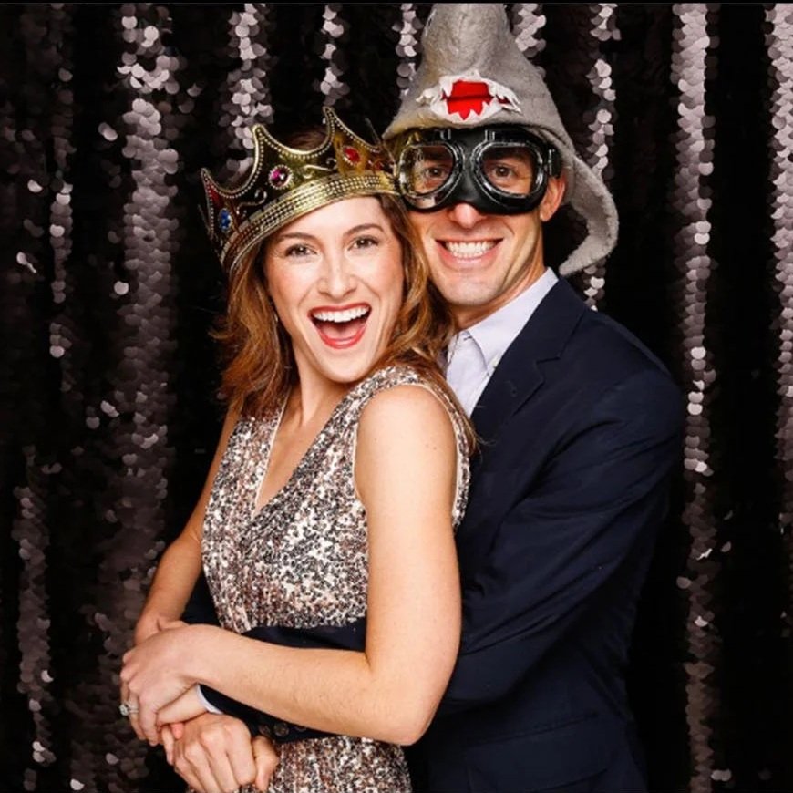 Couple dressed up with party accessories, the woman wearing a sparkling dress and a crown, the man wearing a hoodie hat and goggles, posing joyfully in front of a black sequin backdrop.