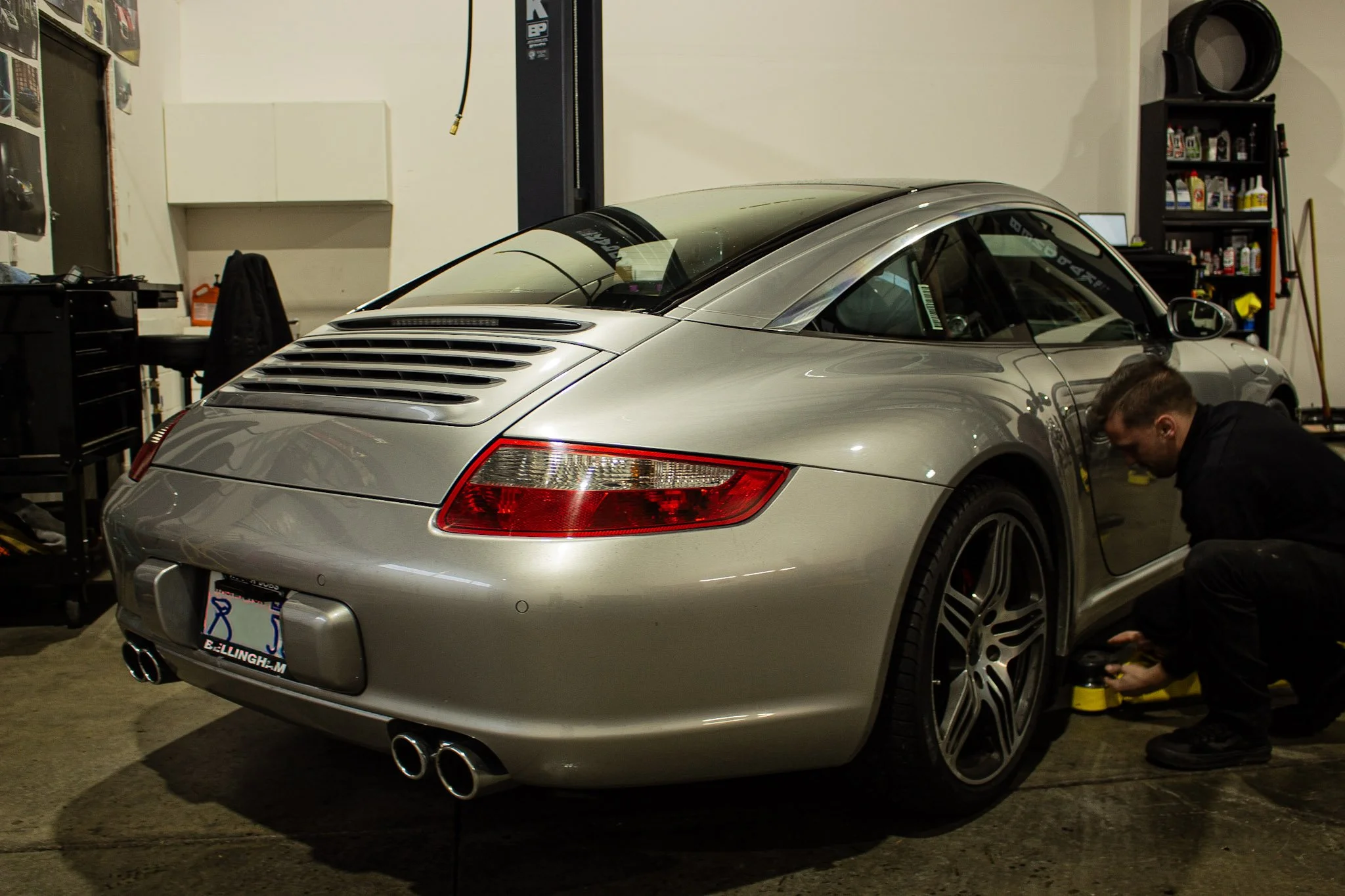 A silver Porsche 911 Targa inside a garage, with a person polishing or working on the front right wheel.