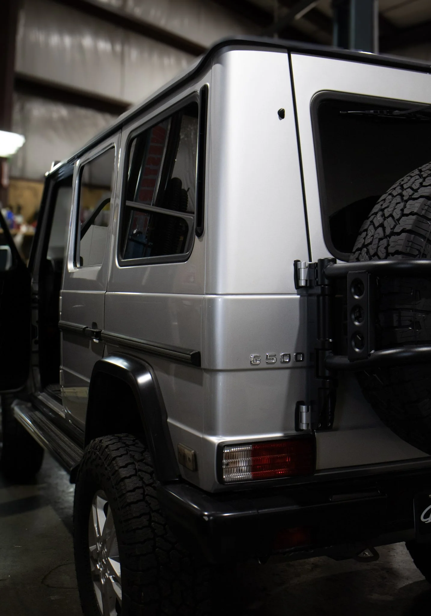 Close-up of the rear side of a silver Mercedes-Benz G-Class SUV, showing part of the rear wheel, rear window, and side panel, inside a garage.