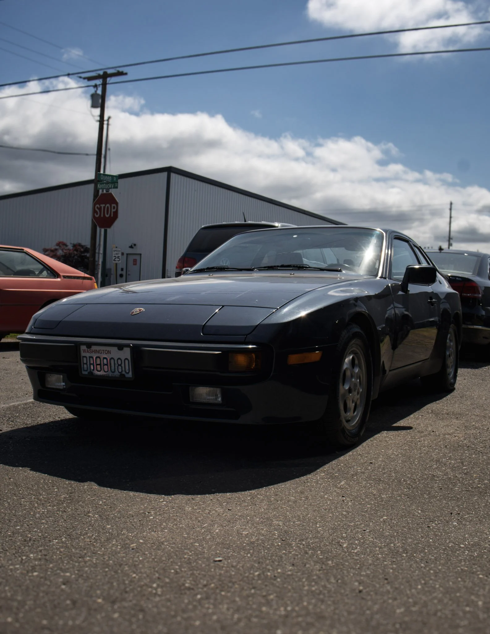 A black Porsche sports car parked on a gravel lot, with other cars and a warehouse building in the background, under a partly cloudy sky.