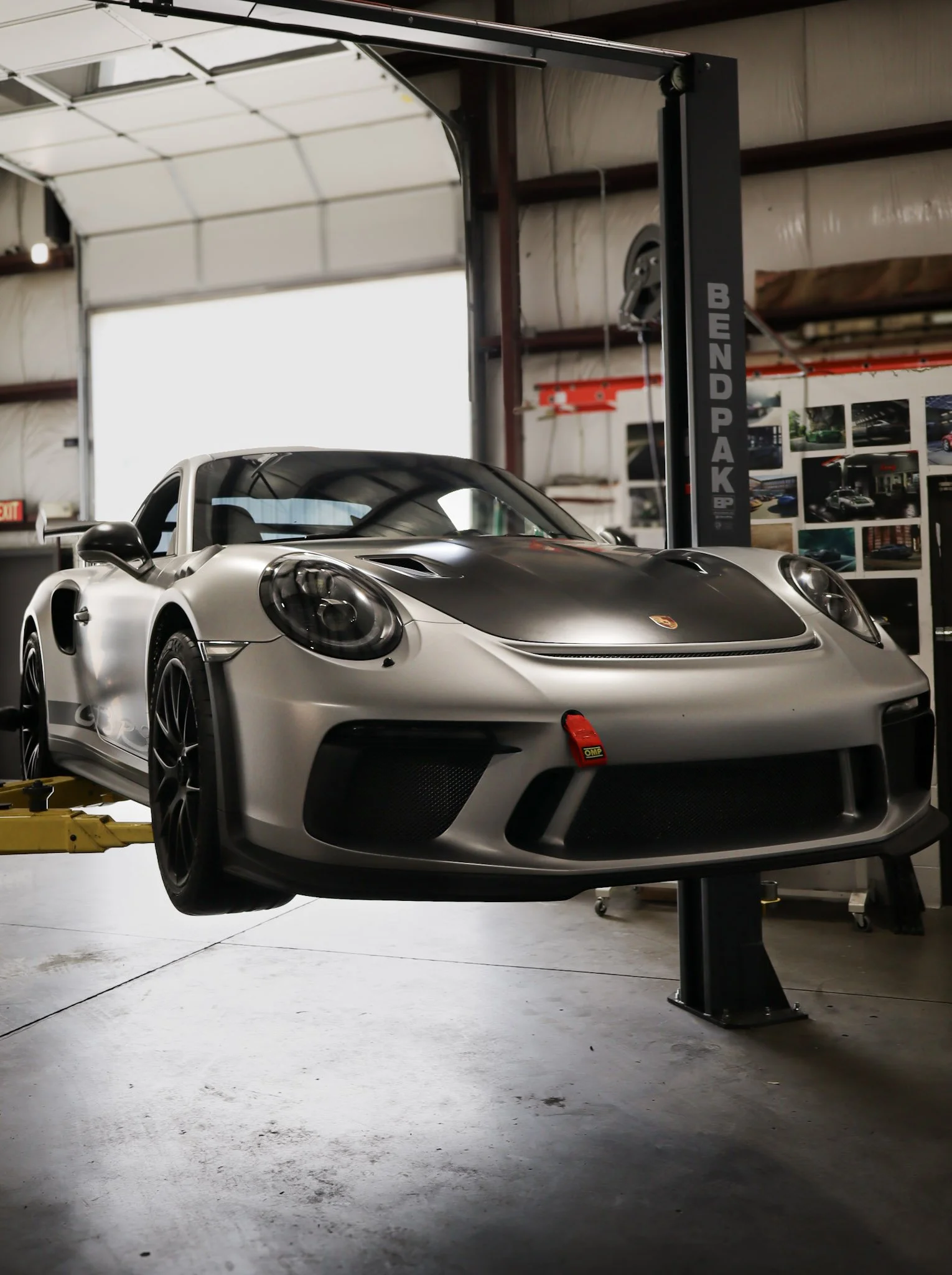 A silver Porsche 918 Spyder race car in a garage on a hydraulic lift with photographs of other cars on the wall in the background.
