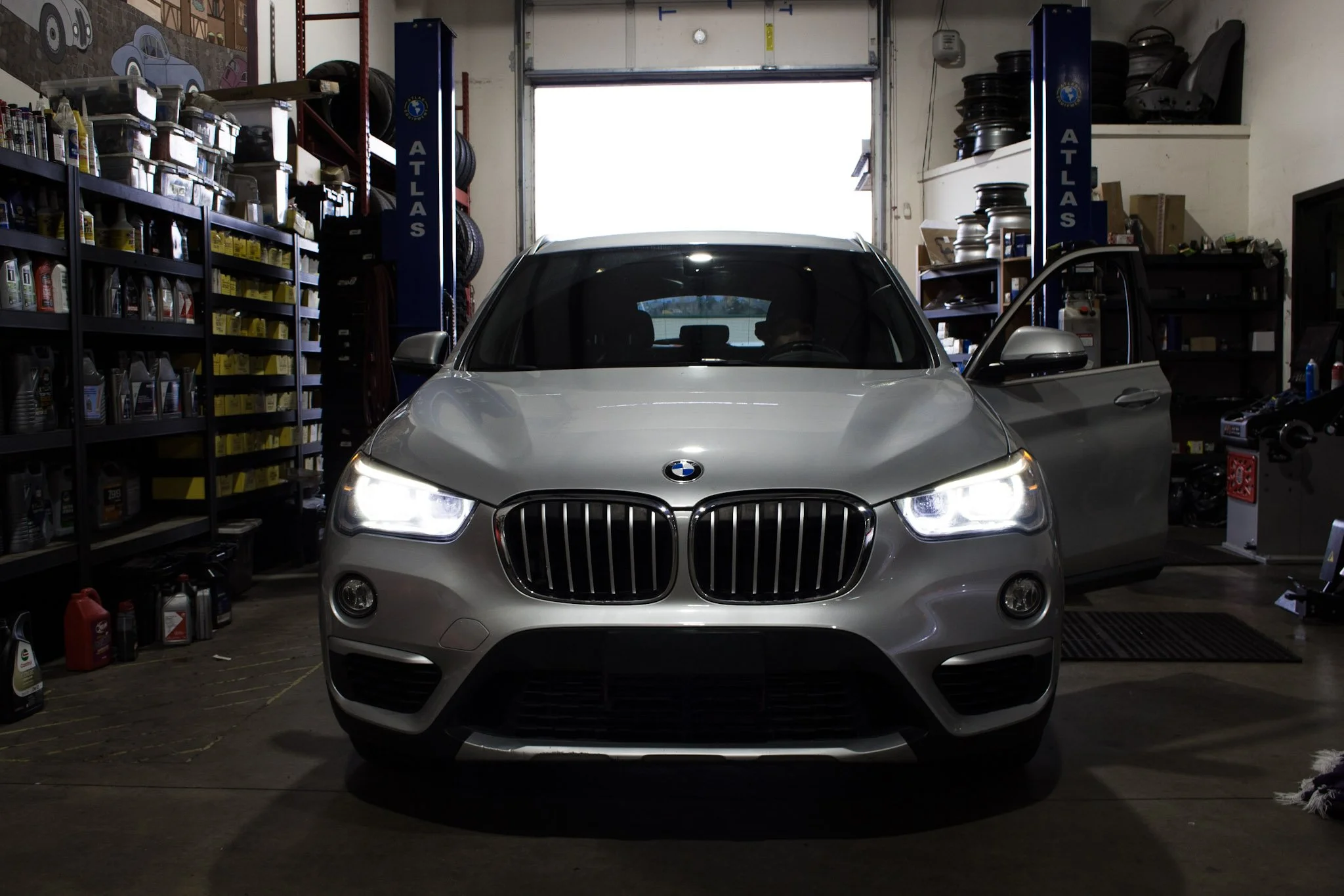 A silver BMW SUV inside a garage or auto repair shop with the driver's side door open. Shelves with car supplies and tires are visible around the space.