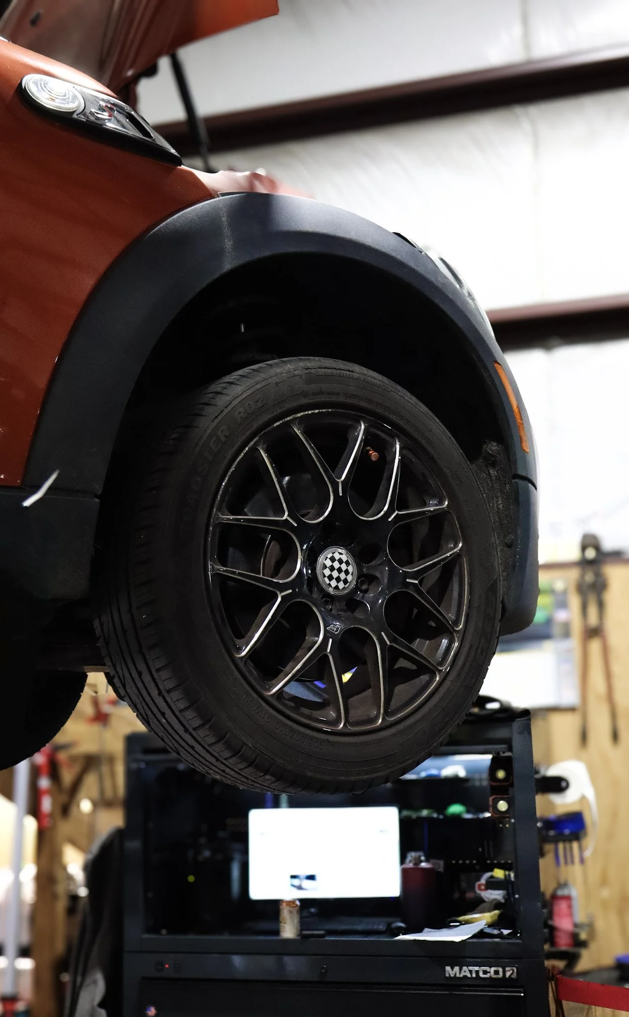 Close-up of a car wheel mounted on a rim, elevated on a lift in a garage or workshop with tools and equipment in the background.